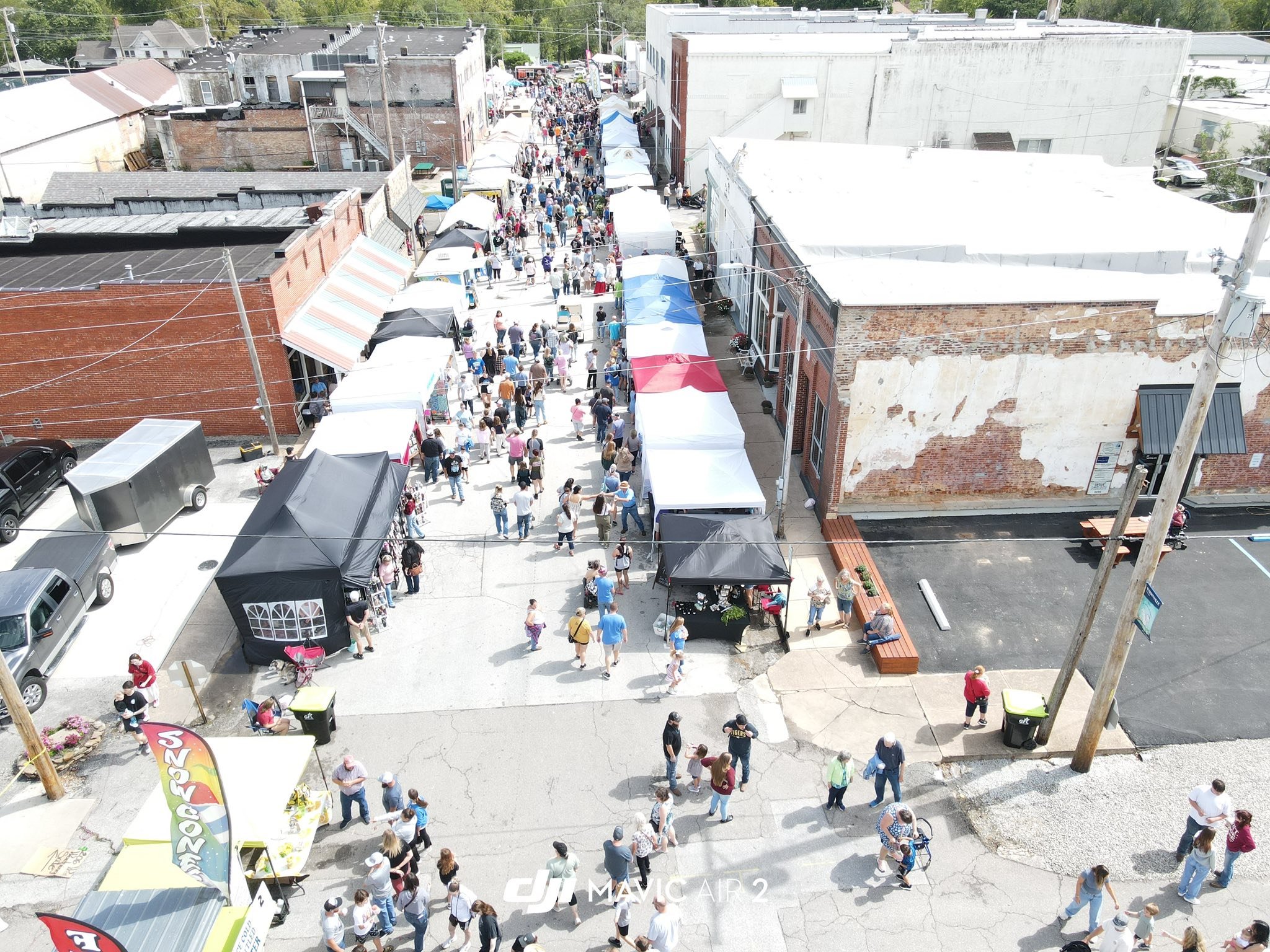 Overhead view of a busy outdoor street fair with tents and stalls, crowded with people walking along the street and sidewalks, surrounded by old brick buildings and parked cars.