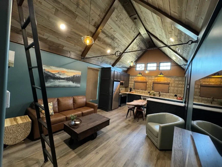 Cozy living space with a leather sofa, a white armchair, a wooden coffee table, and a modern kitchen area with stainless steel appliances and a patterned backsplash.