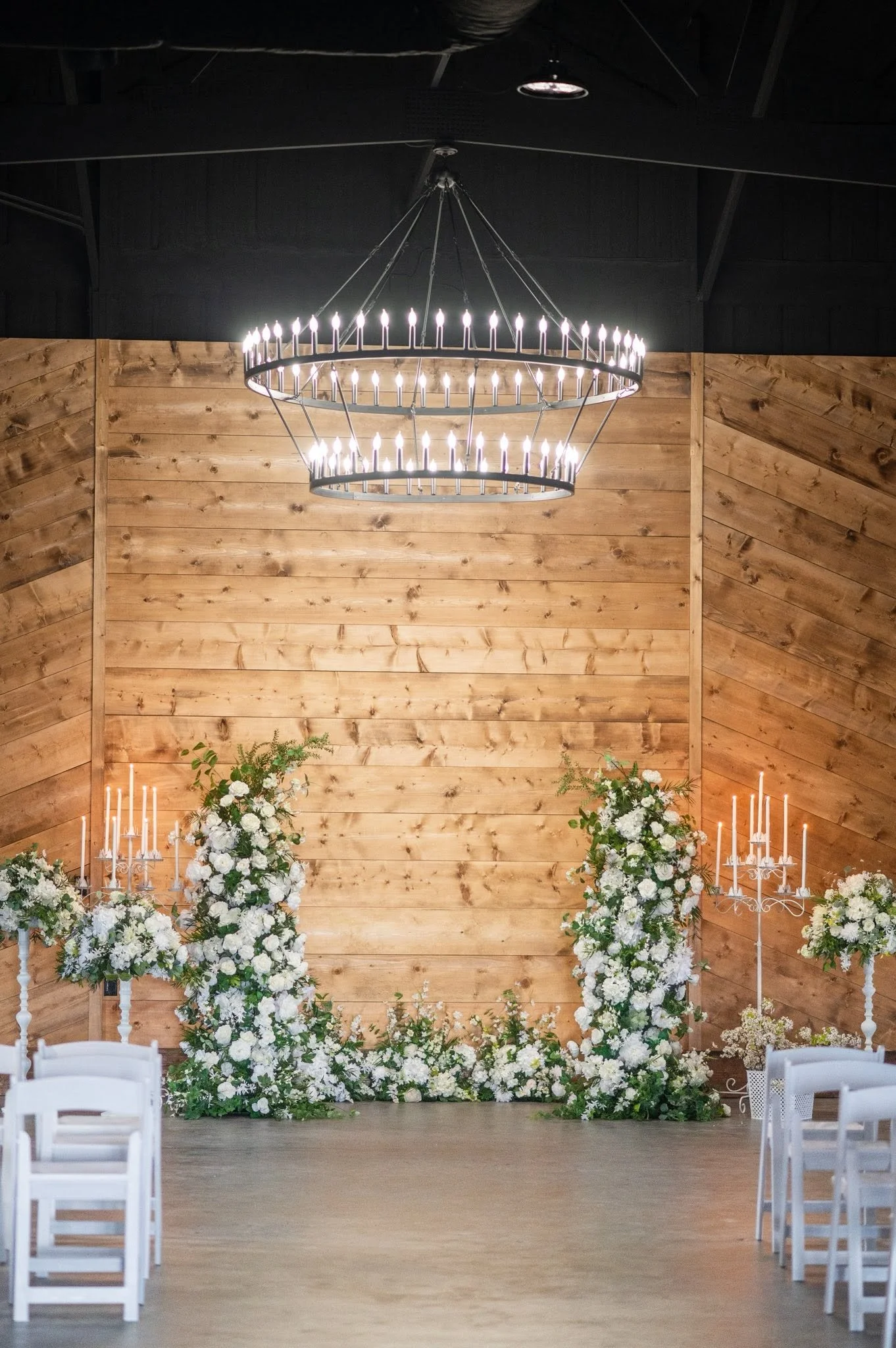 Indoor wedding venue with wooden walls, large floral arrangements, and a circular chandelier with candle-shaped lights hanging from the ceiling.