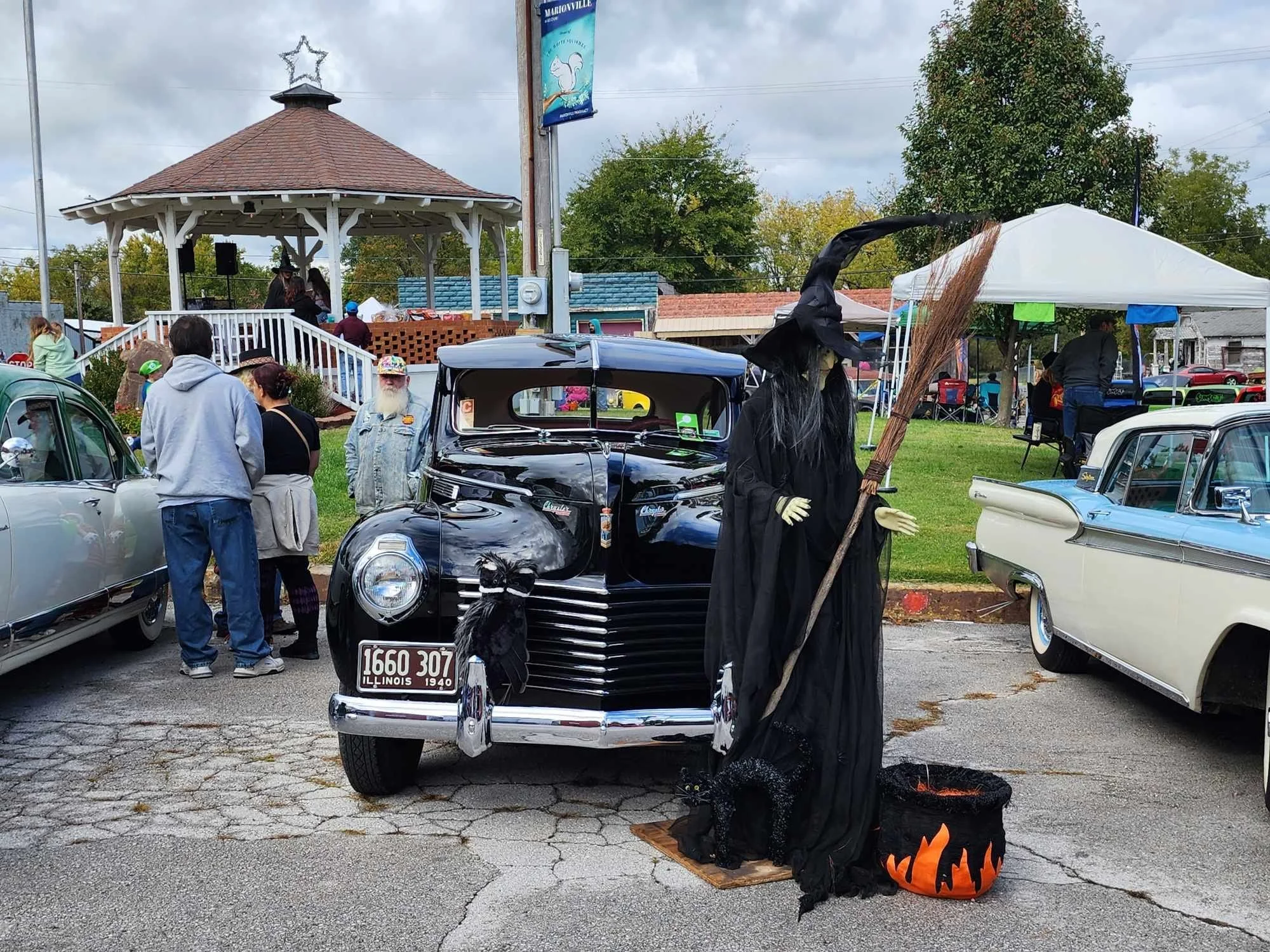 A Witch decoration standing in front of a vintage black car at a community event or car show, with people gathered around and a gazebo with an American flag banner in the background.