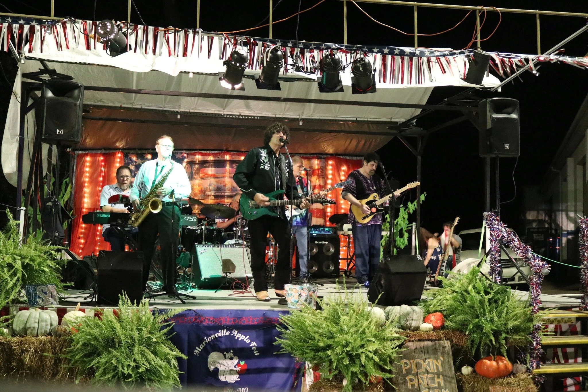 A band performing on a decorated outdoor stage at night, with five musicians playing various instruments and a backdrop with festive lights. The stage is adorned with American-themed decorations and pumpkins, indicating a fall or harvest festival.