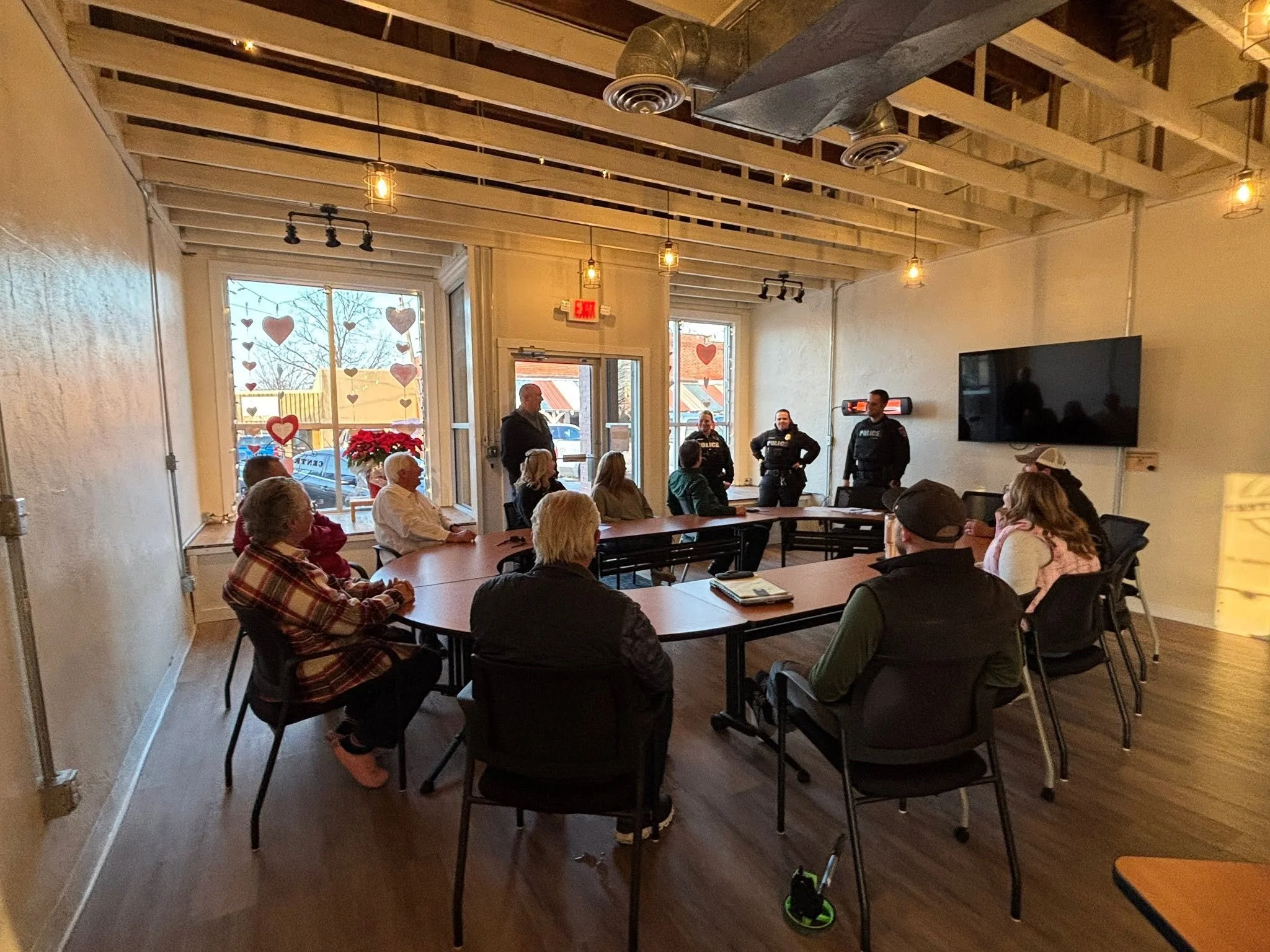A group of people, including police officers, sitting around a large table listening to a speaker inside a room decorated with Valentine's Day hearts and lights, with large windows showing trees outside.