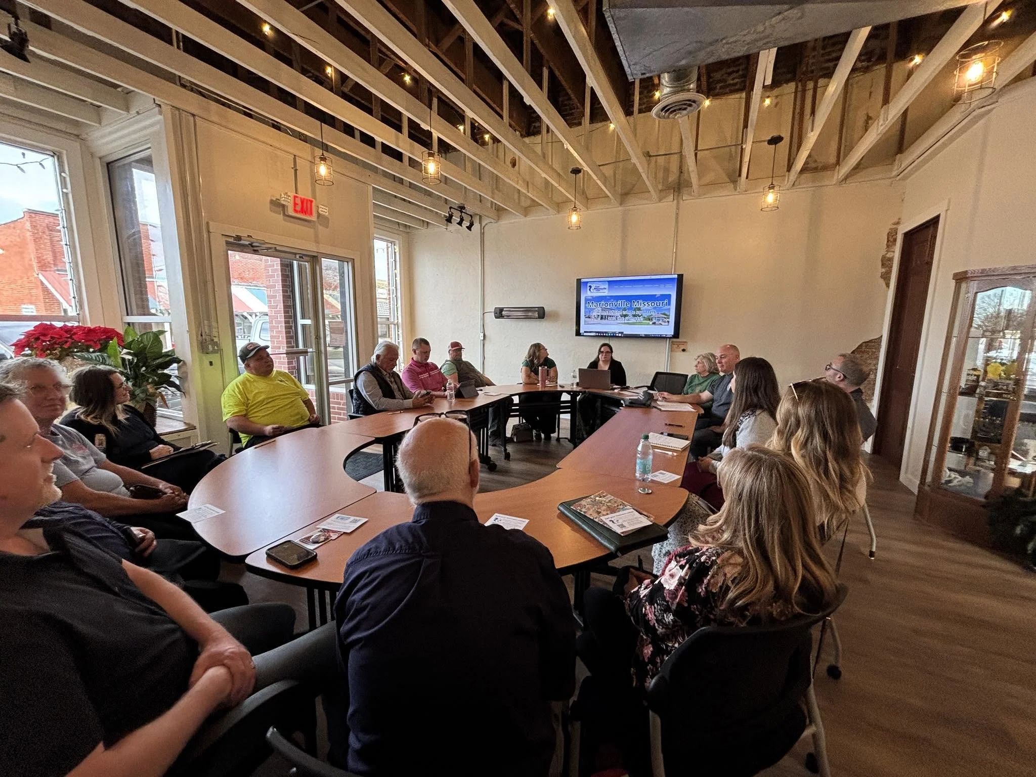 People sitting around a large conference table in a room with high ceilings, exposed beams, and large windows. There is a TV screen displaying 'Marionville Missouri' on a wall, and a poinsettia plant is visible near the window.