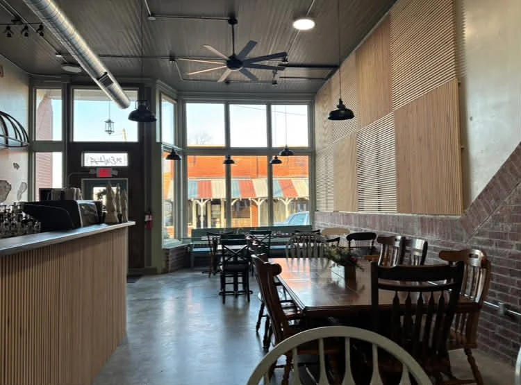 Interior of a cozy restaurant or coffee shop with wooden tables and chairs, large front windows, hanging pendant lights, a ceiling fan, and a brick accent wall.