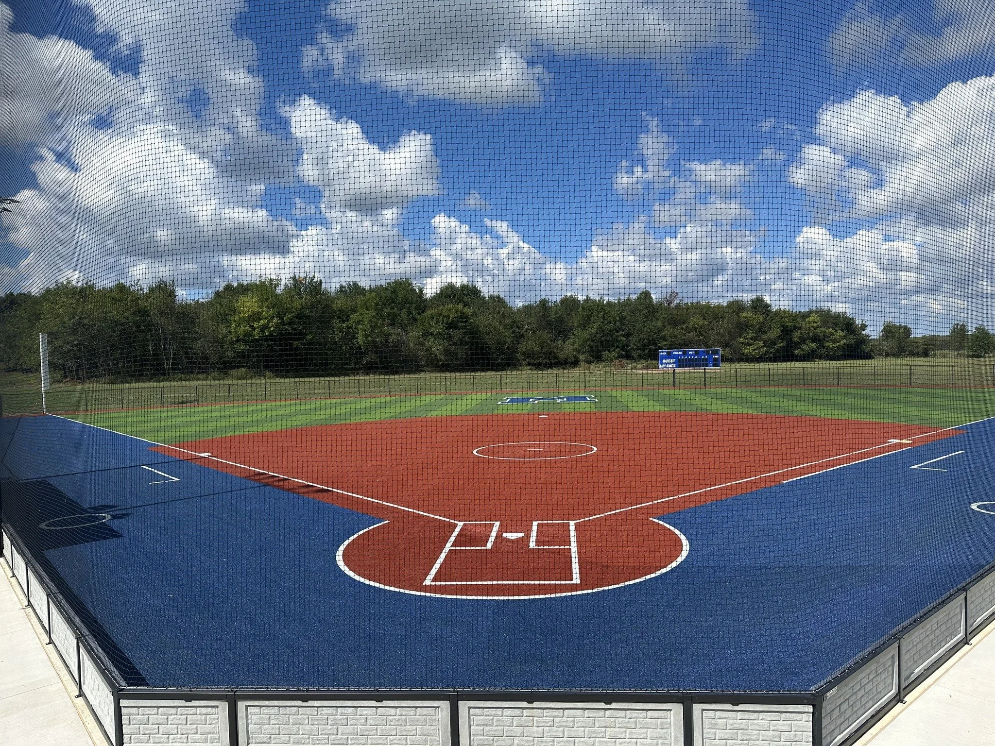 Empty baseball field with a red infield, green outfield, blue warning track, and a scoreboard in the distance under a partly cloudy sky.