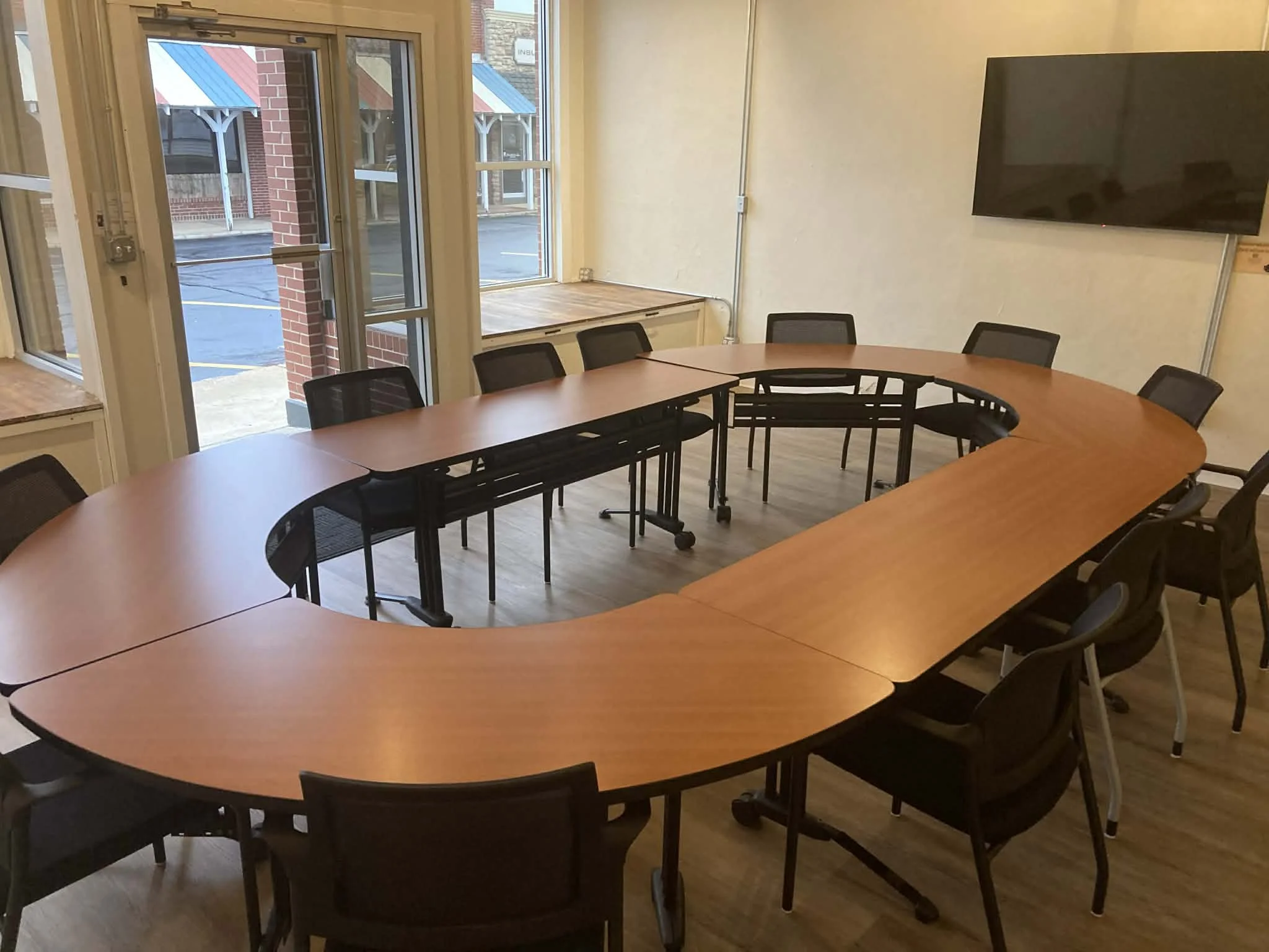 Meeting room with a U-shaped table arrangement, black chairs, large windows, and a wall-mounted TV.