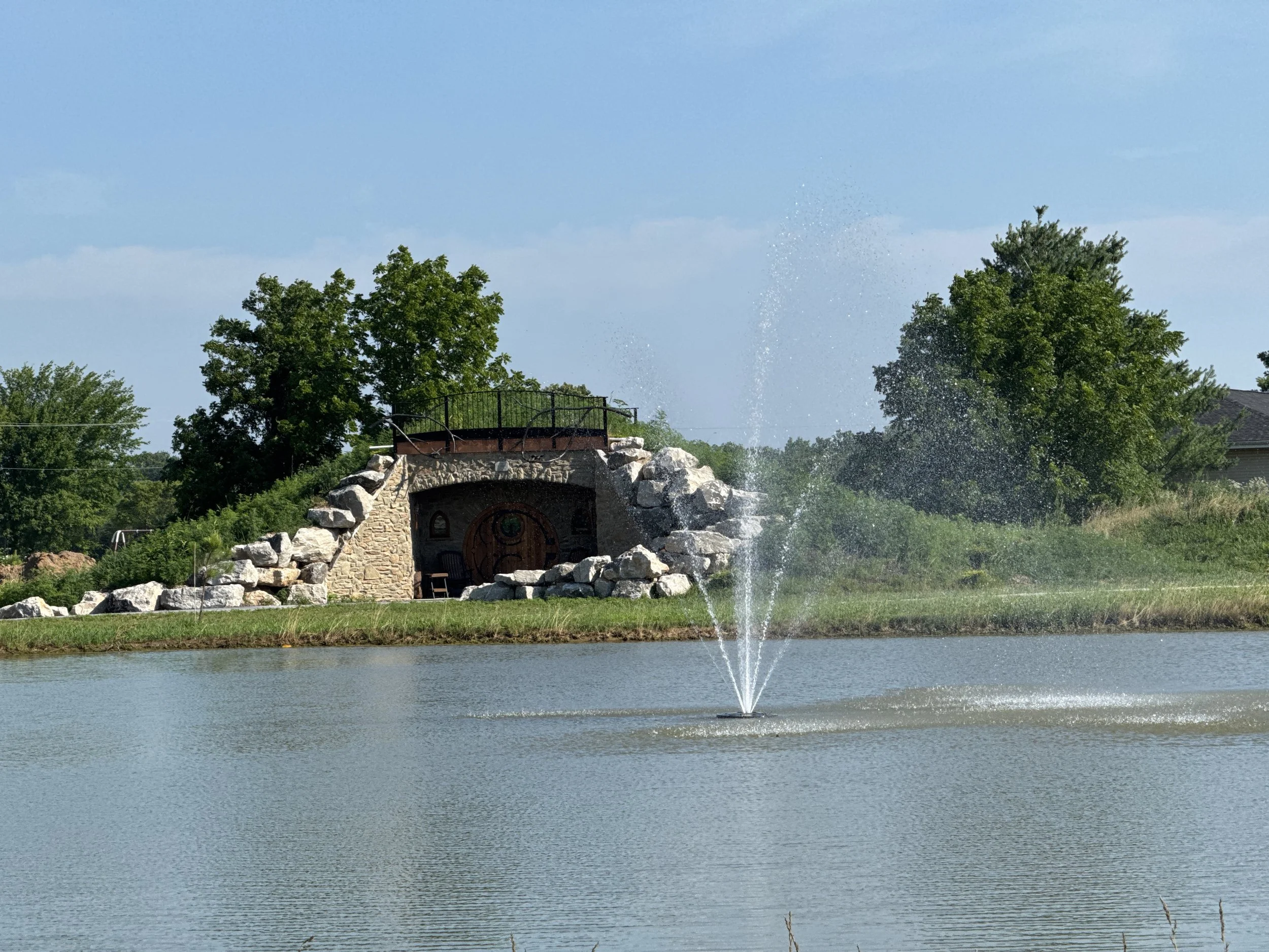 View of a pond with a water fountain, a stone structure with a wooden door and a balcony behind it, surrounded by trees and greenery.