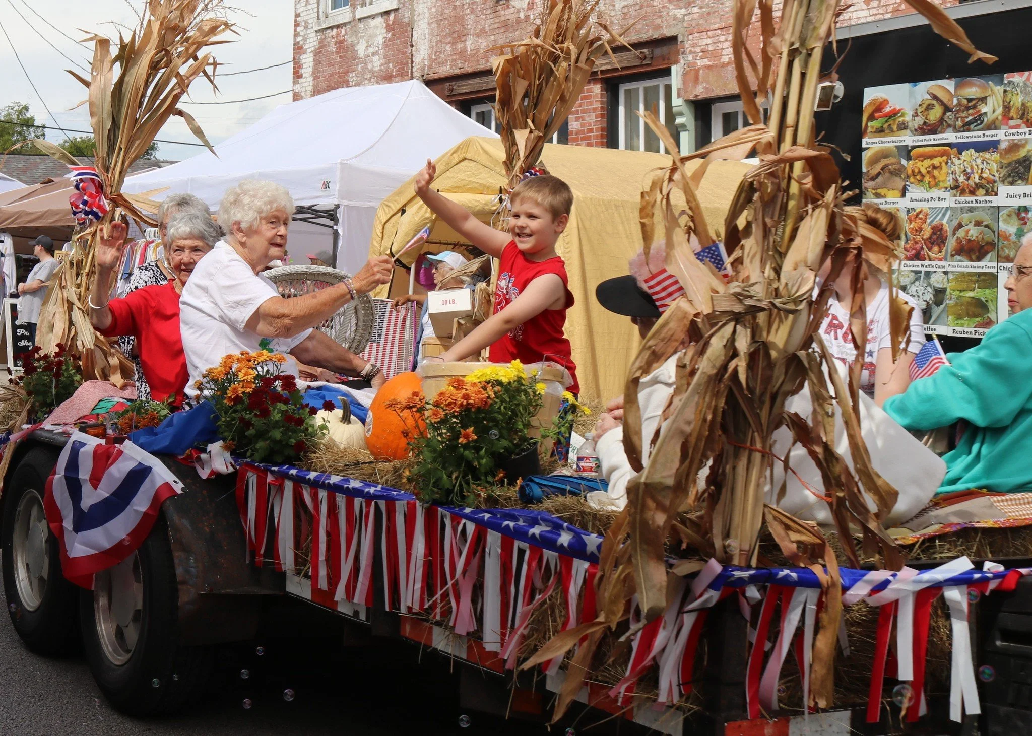 A parade float decorated with American flags, corn stalks, and pumpkins, featuring elderly women and children celebrating during a patriotic festival or parade, with tents and a food stand in the background.