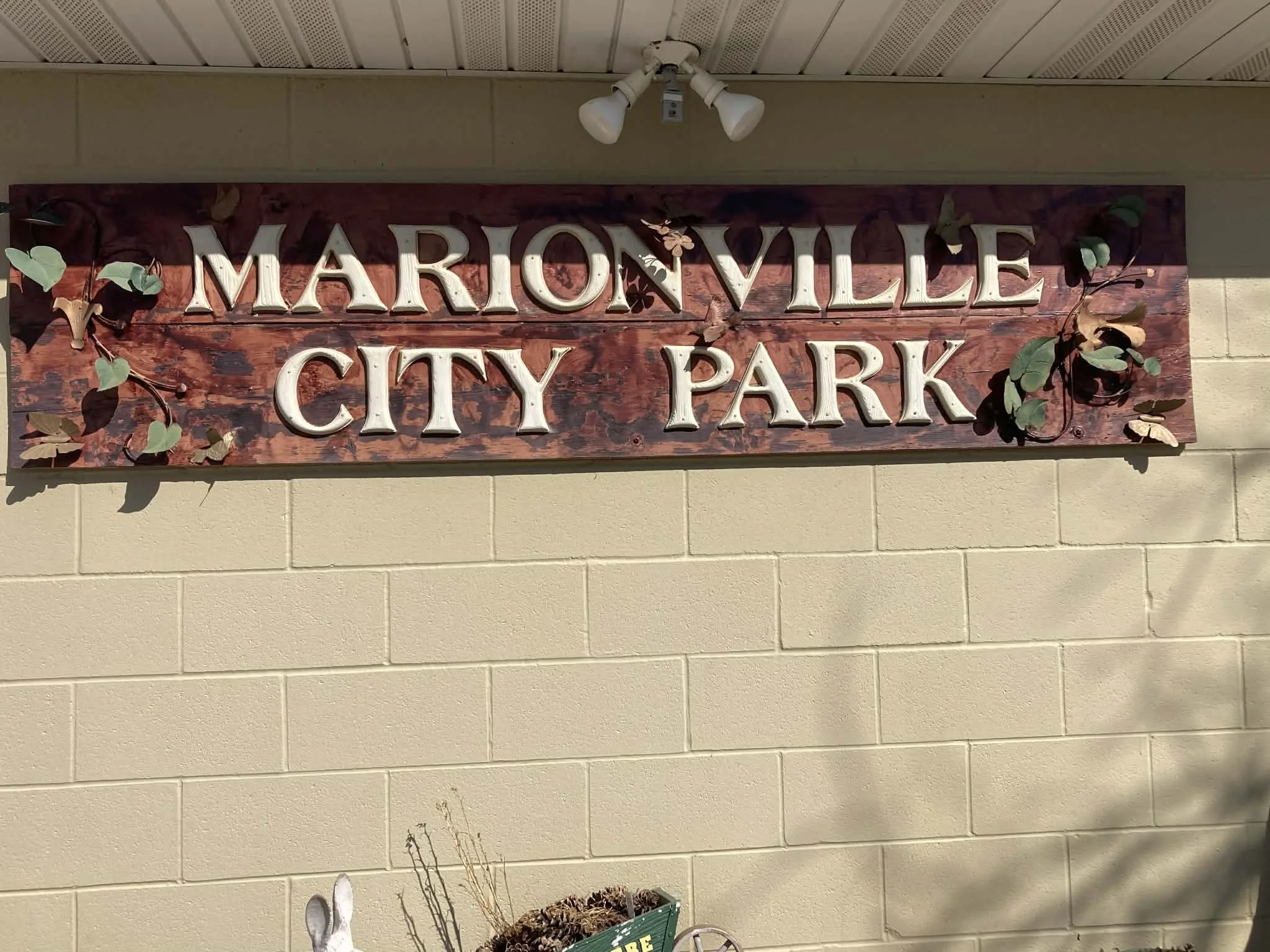 Wooden sign reading 'Marionville City Park' with decorative vines and leaves, mounted on beige brick wall.