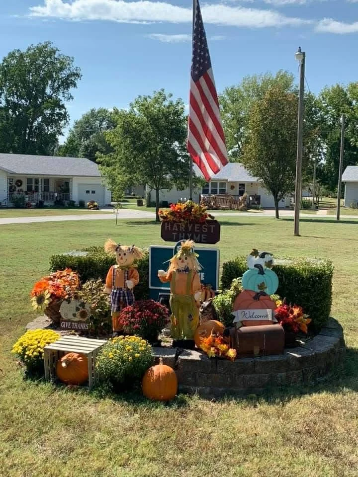 Fall harvest display with pumpkins, gourds, flowers, scarecrows, a decorated scarecrow with a plaid shirt, and an American flag on a flagpole in a grassy yard with houses and trees in the background.
