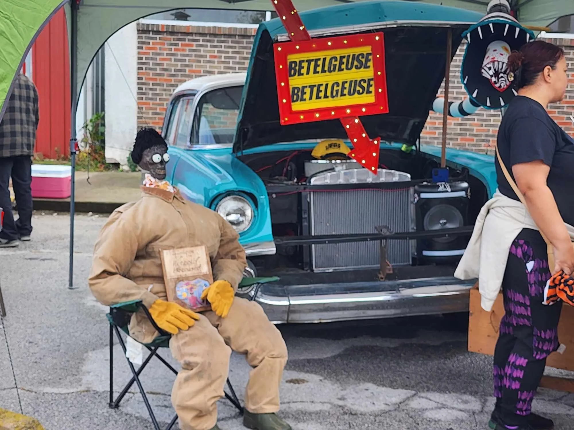 A vintage blue car with its hood open, displaying a sign reading 'BETELGEUSE'. A skeleton figure wearing a tan coverall and yellow gloves sits on a folding chair beside the car, holding a small colorful item. There are people nearby, one of whom is d