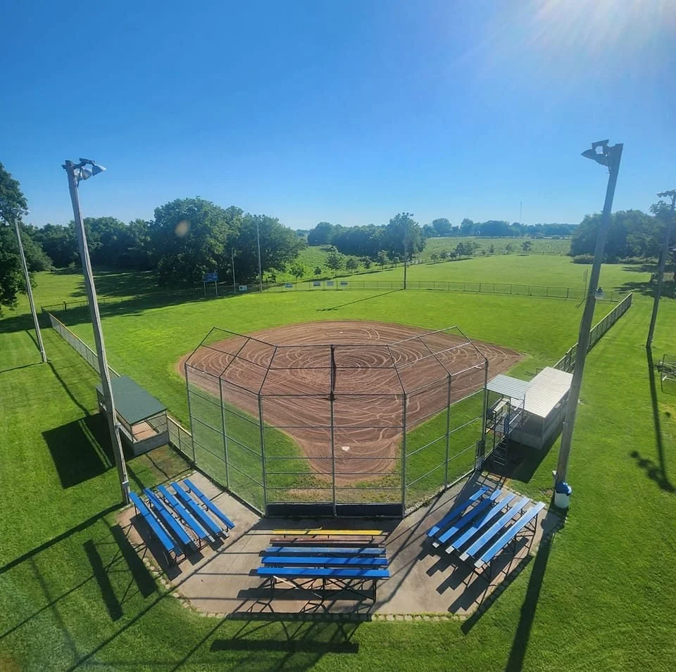 An aerial view of a baseball field with a dirt infield, surrounded by green grass and open fields, under a clear blue sky with sunlight.