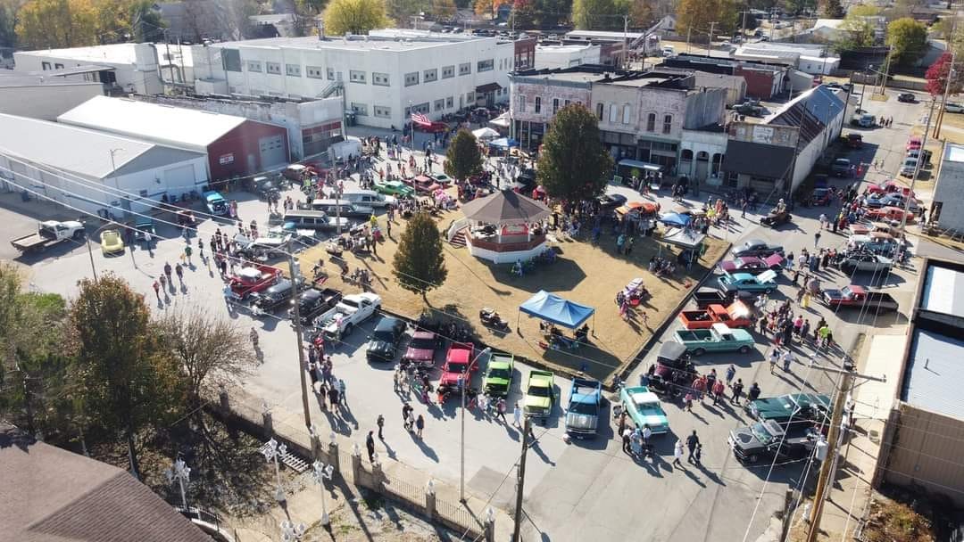 A lively outdoor event with a crowd of people gathered around a small park with a gazebo, surrounded by vintage cars and vendor tents, in a downtown area with buildings and parking lots.