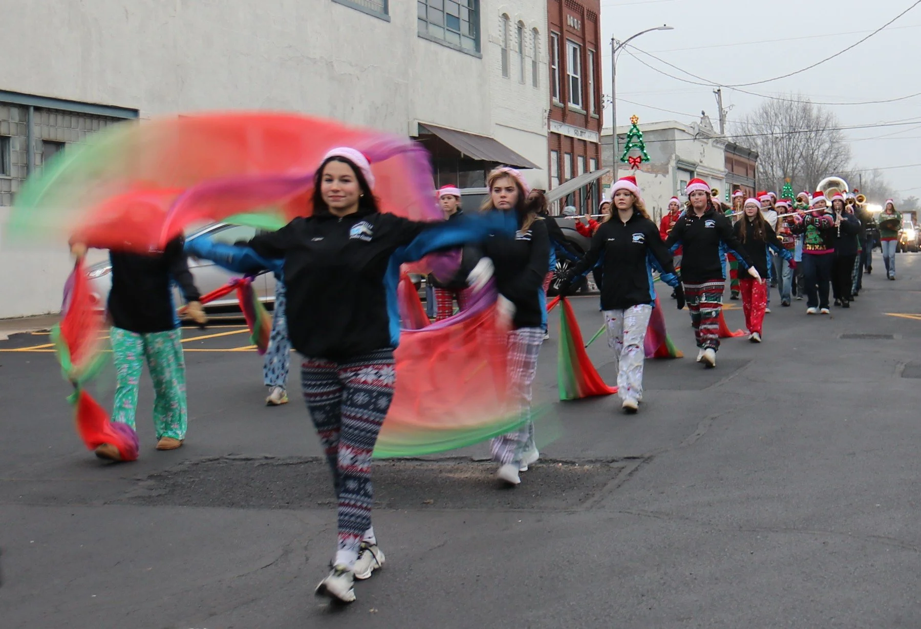 A parade of young women dressed in Christmas pajamas and jackets, some wearing Santa hats, marching down a street with festive decorations including Christmas trees, and a band playing instruments in the background.