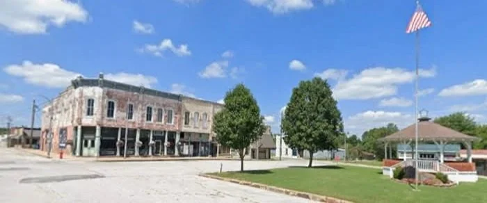 A small town square with a historic brick building, a flagpole with an American flag, and a gazebo surrounded by trees and a lawn.