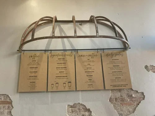 Menu board with coffee categories and drinks, hanging under a metal rack on a wall with exposed brick.