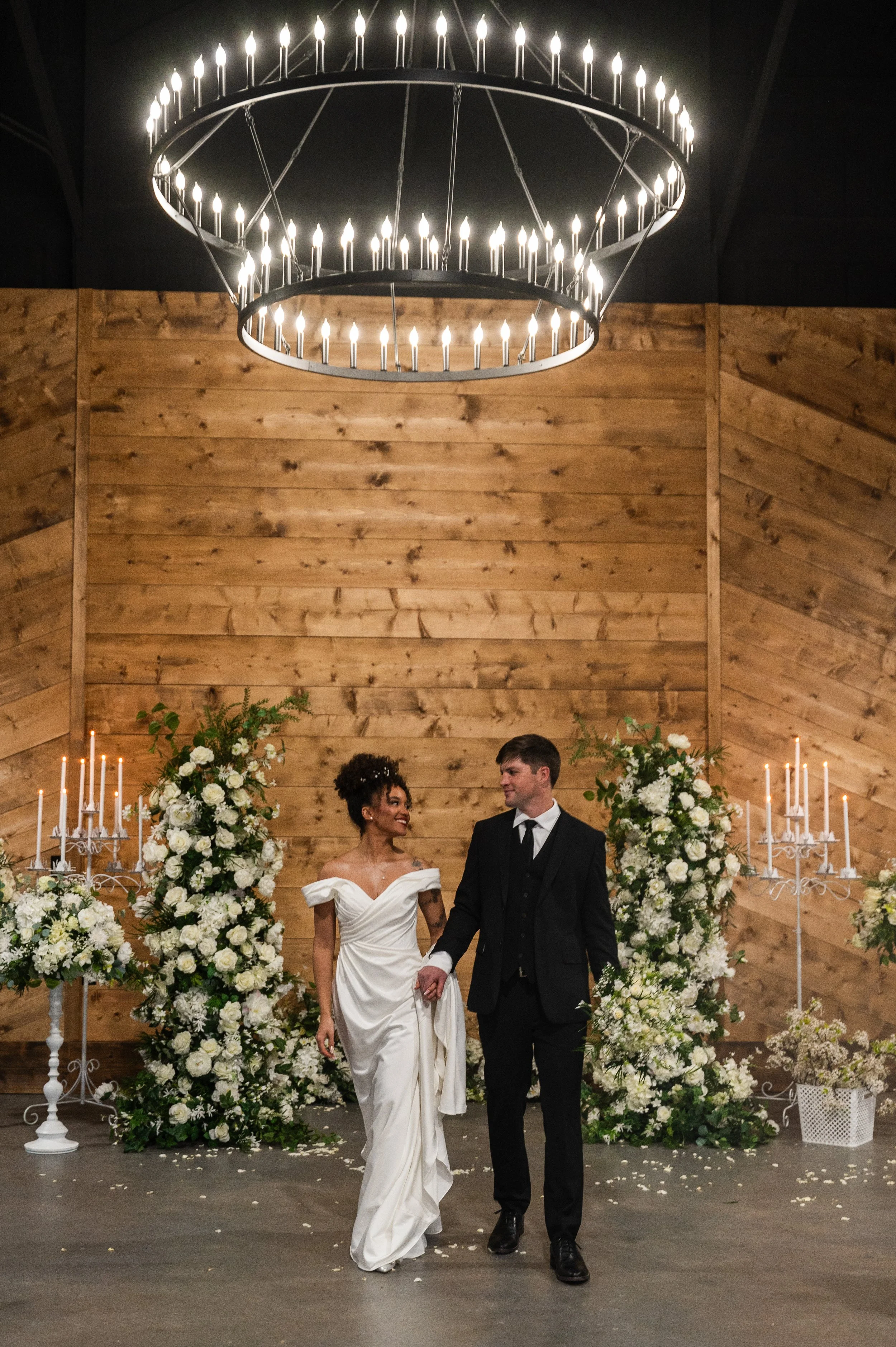 A bride and groom holding hands and walking in a wedding ceremony, surrounded by large floral arrangements with white flowers, in a barn-style venue with wooden walls and a circular chandelier with lit candles hanging from the ceiling.