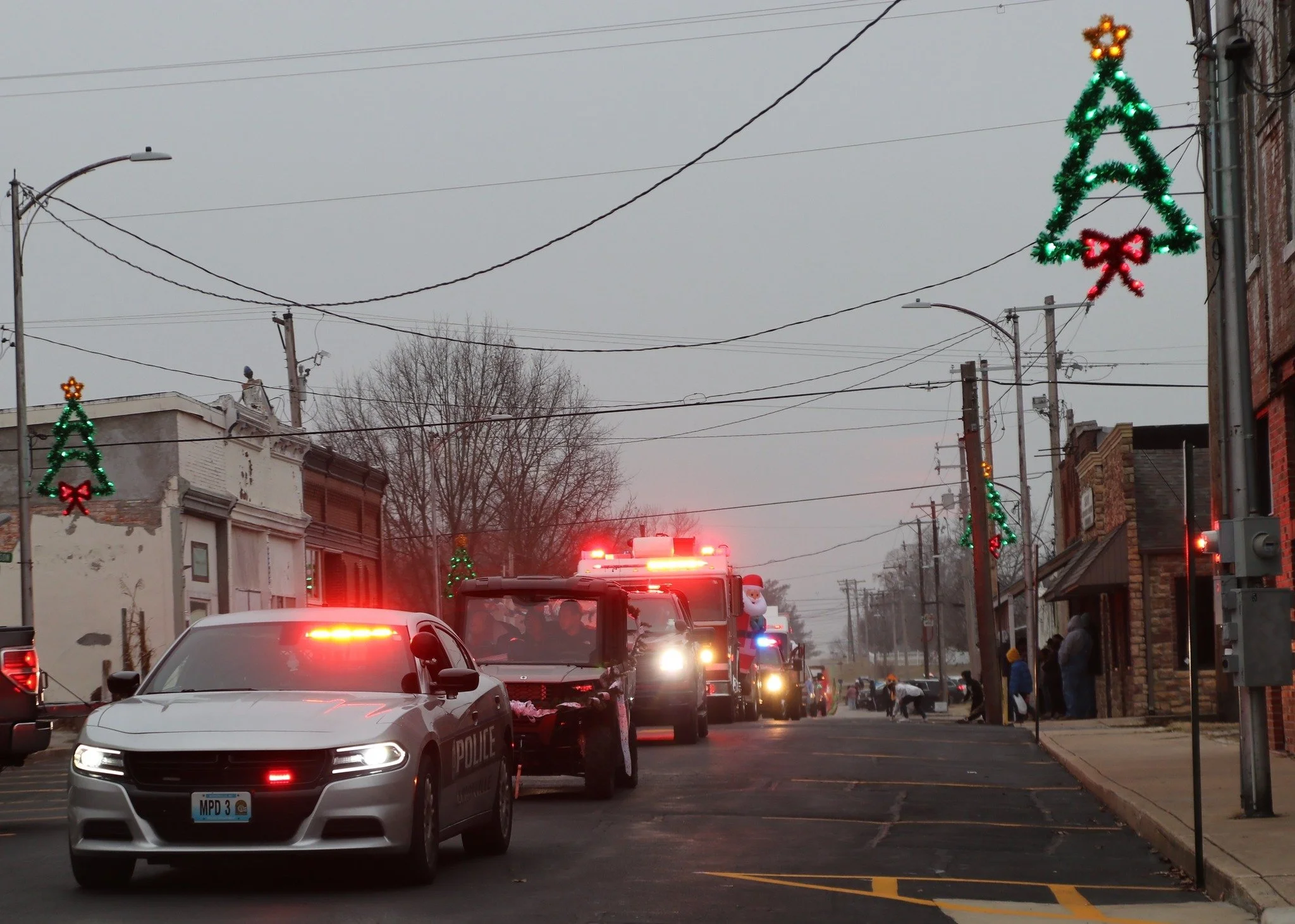 A winter street scene decorated with Christmas lights and ornaments, with police and emergency vehicles present.