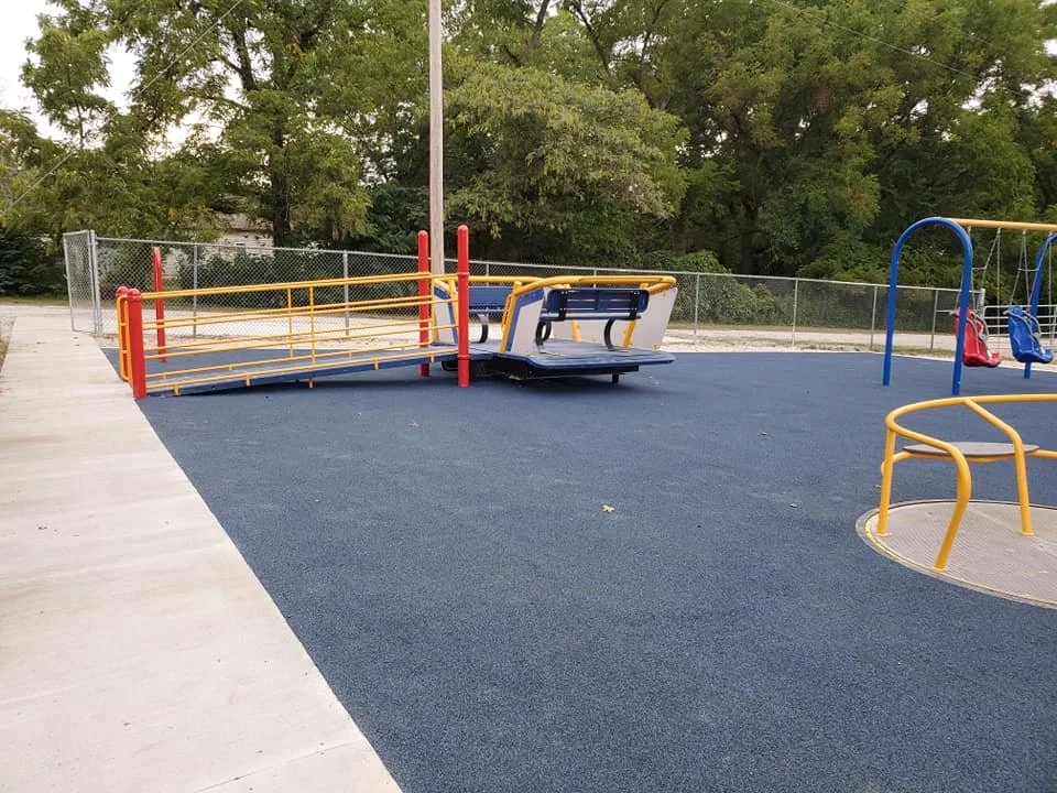 Empty playground with blue rubberized flooring, metal railings, and swings set in the background, surrounded by trees.
