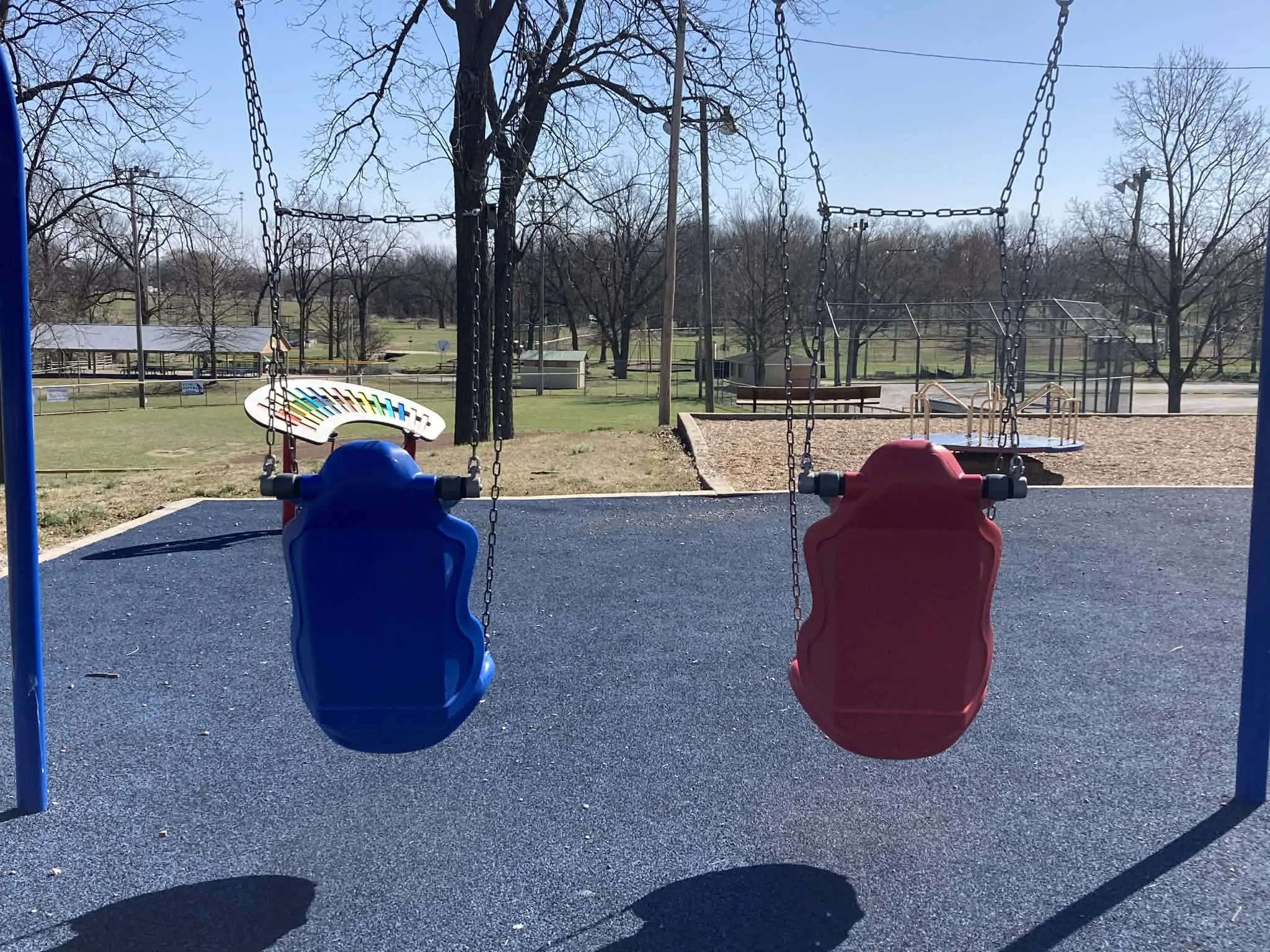 Empty inclusive swing set with blue and red swings at a playground, with a field and trees in the background. At Marionville MO city park.