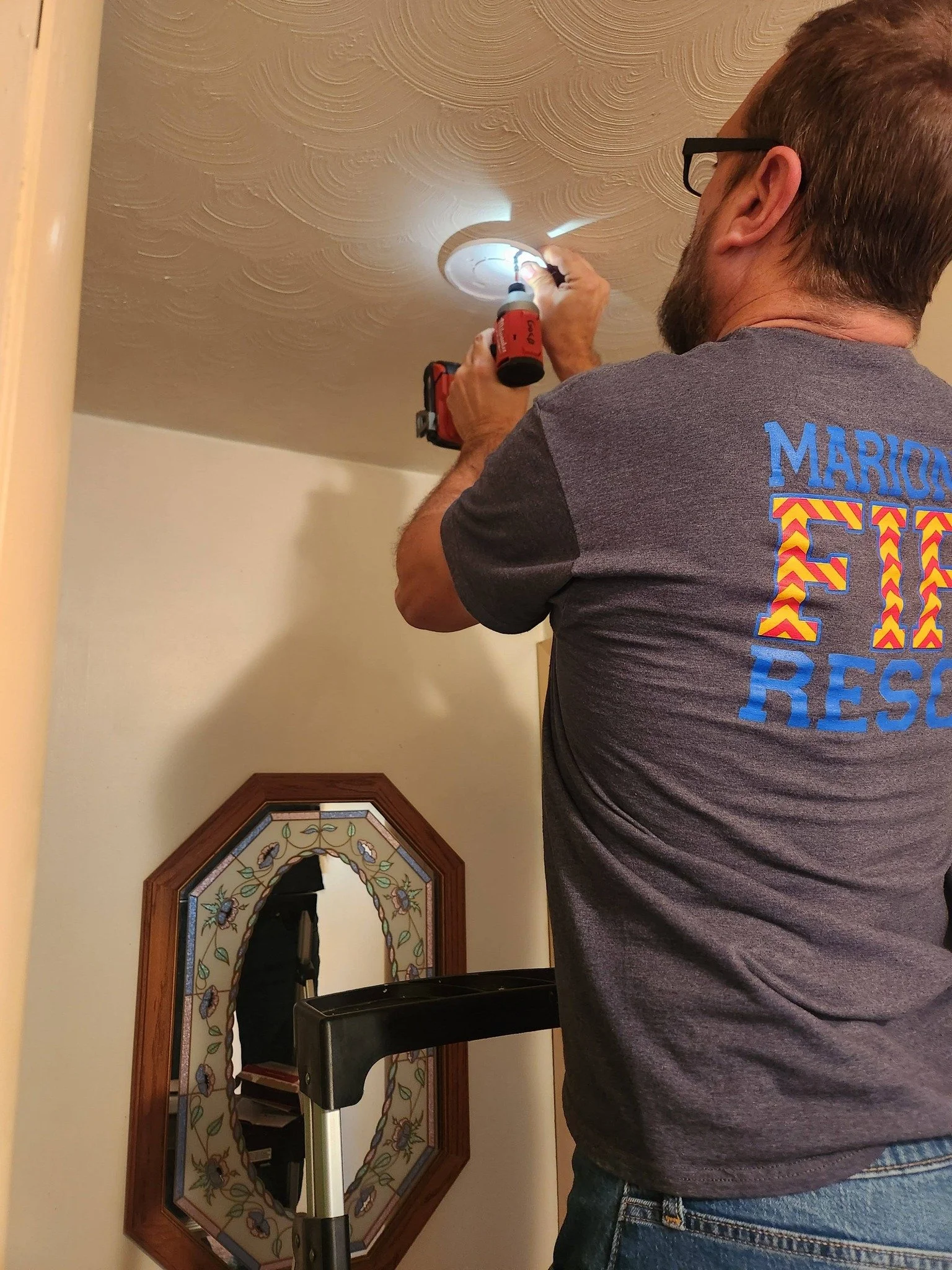 A man with glasses, a beard, and a gray shirt is using a screwdriver to work on a ceiling corner near a decorative mirror.