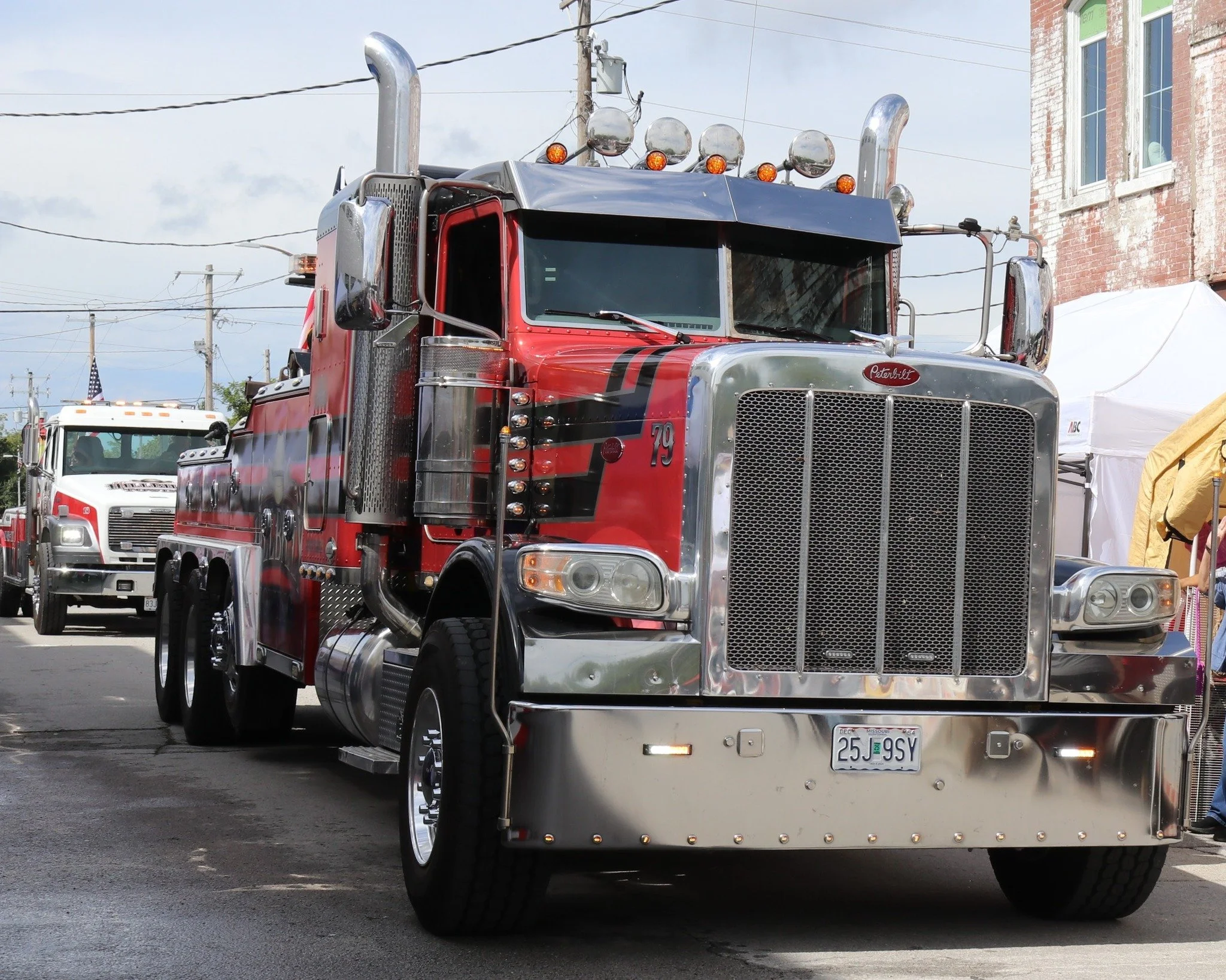 Front view of a red Peterbilt fire truck with chrome accents, parked outdoors during the day, with other emergency vehicles and a tent in the background.