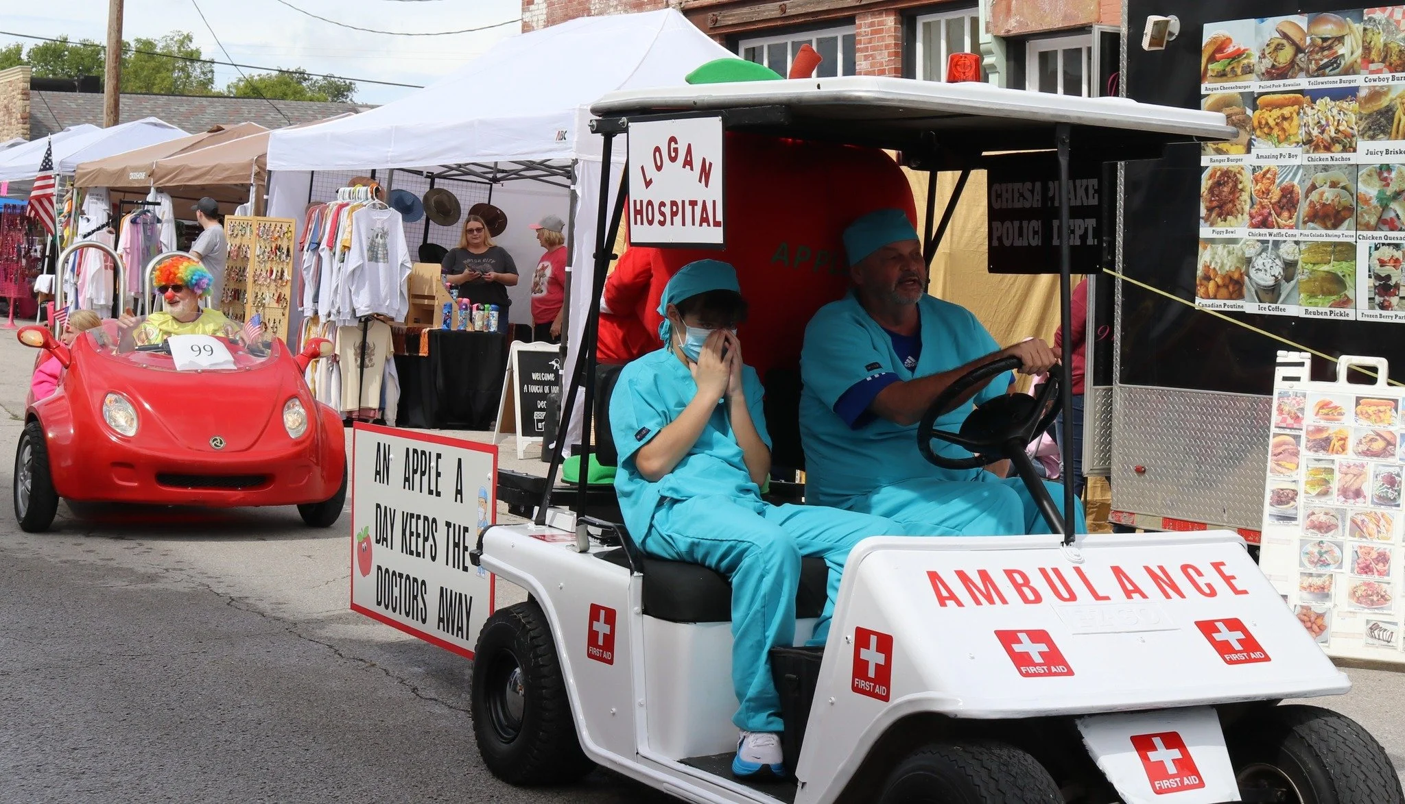 A small parade with a man dressed as a clown riding a toy car, a person dressed as a doctor driving a mini ambulance with a young person in medical scrubs, and vendor booths with clothing and food menus in the background.