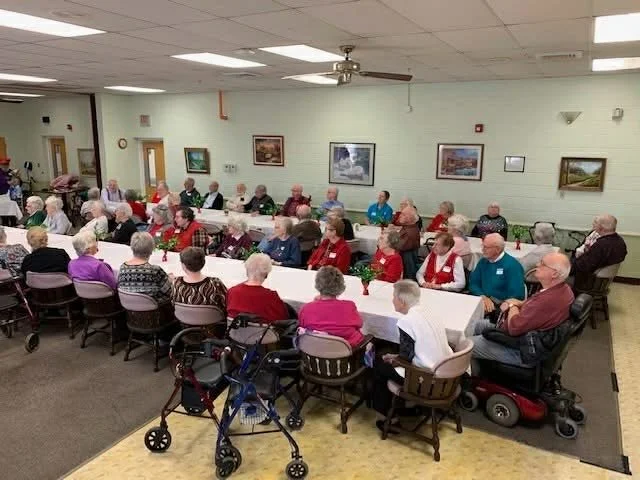 Elderly people gathered at a long rectangular table with holiday decorations in a community room, some using wheelchairs or walkers, with framed landscape paintings on the wall.
