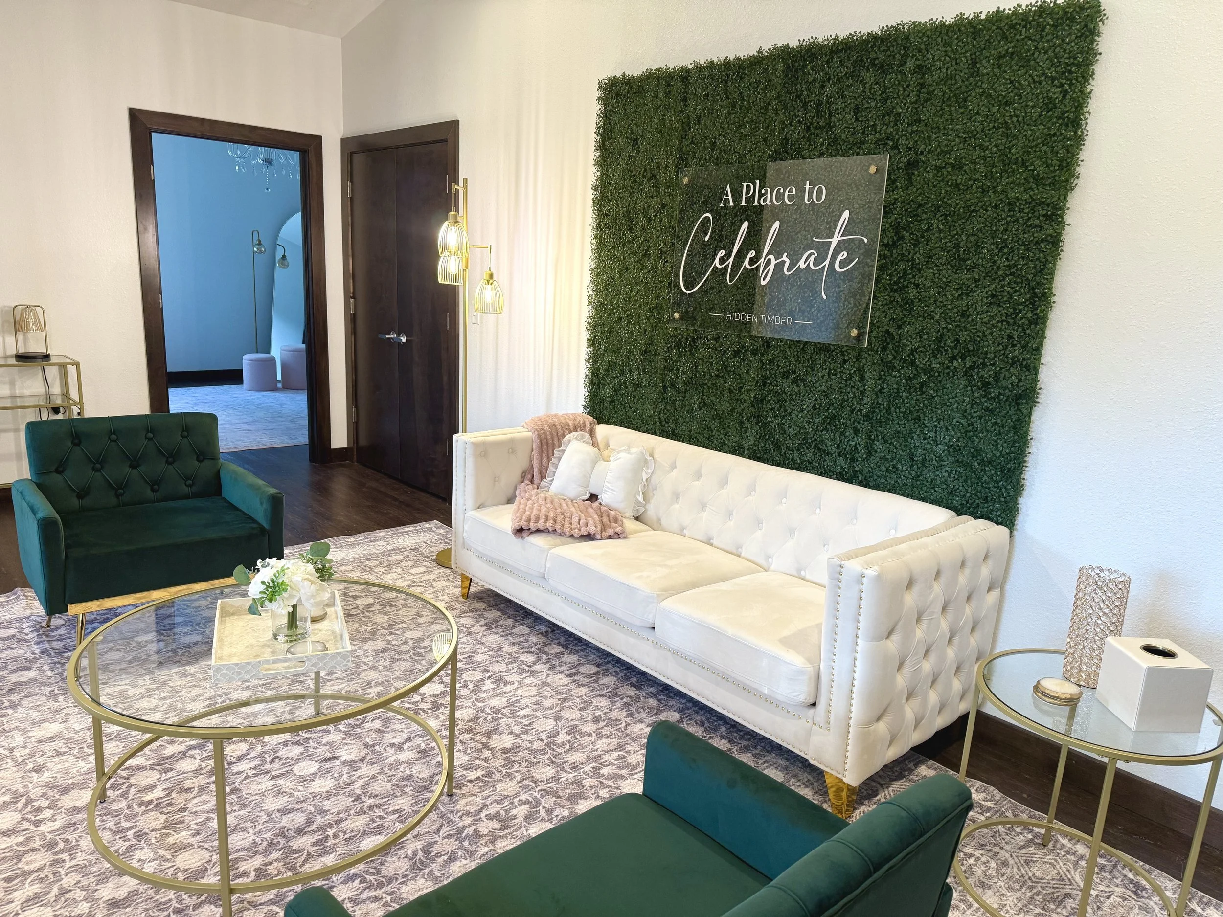 Elegant living room with white tufted sofa, green velvet armchair, gold coffee tables, green wall with sign that reads 'A Place to Celebrate,' decorative pillows and flowers, and doorway leading to another room.