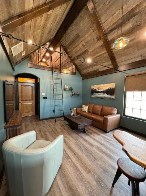 Living room with vaulted wooden ceiling, blue walls, leather sofa, white armchair, wooden coffee table, window with blinds, and wall art