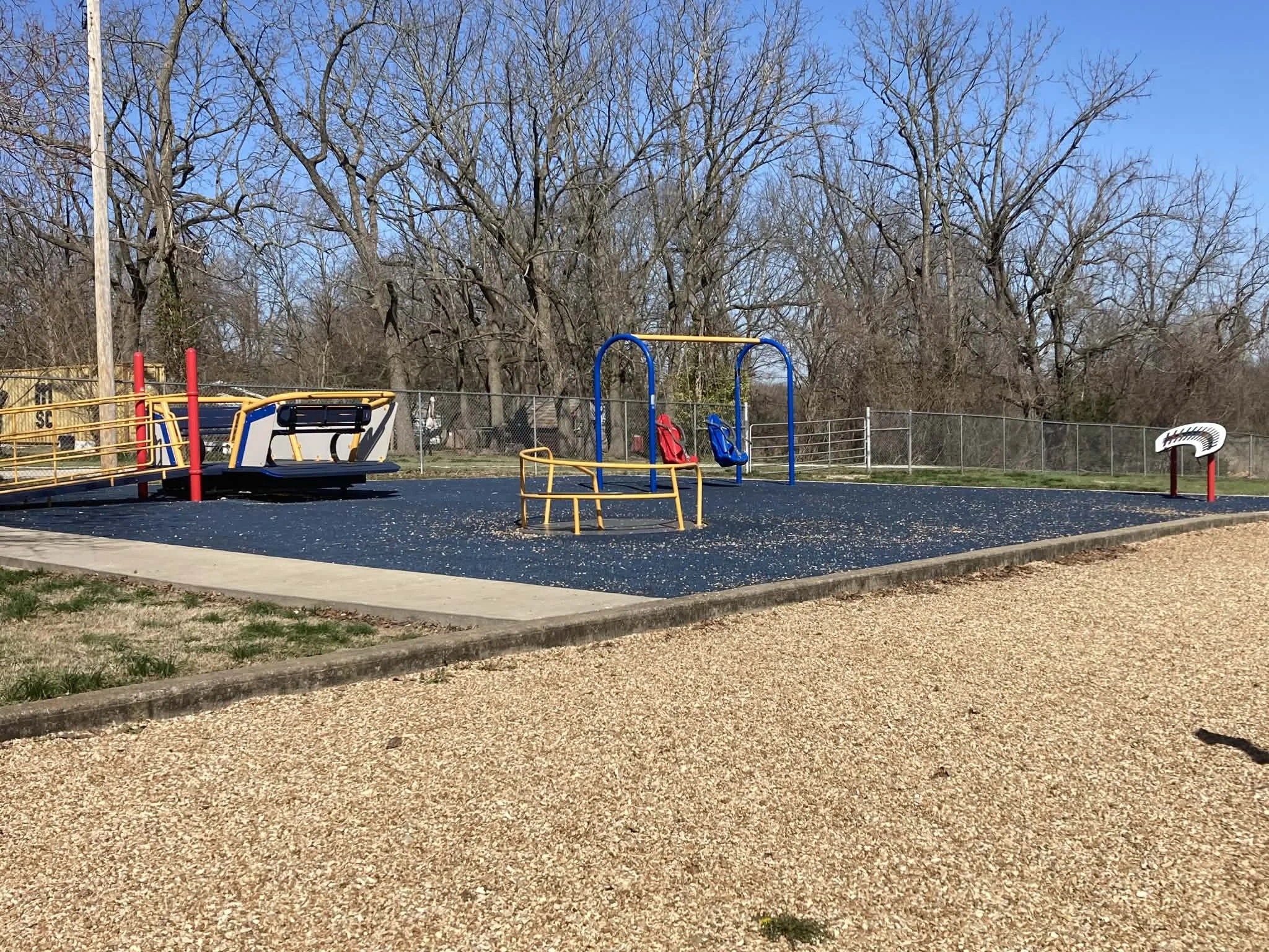 Colorful inclusive playground with swings, a merry-go-round, and a music-themed sculpture, surrounded by a chain-link fence and leafless trees under a clear blue sky.