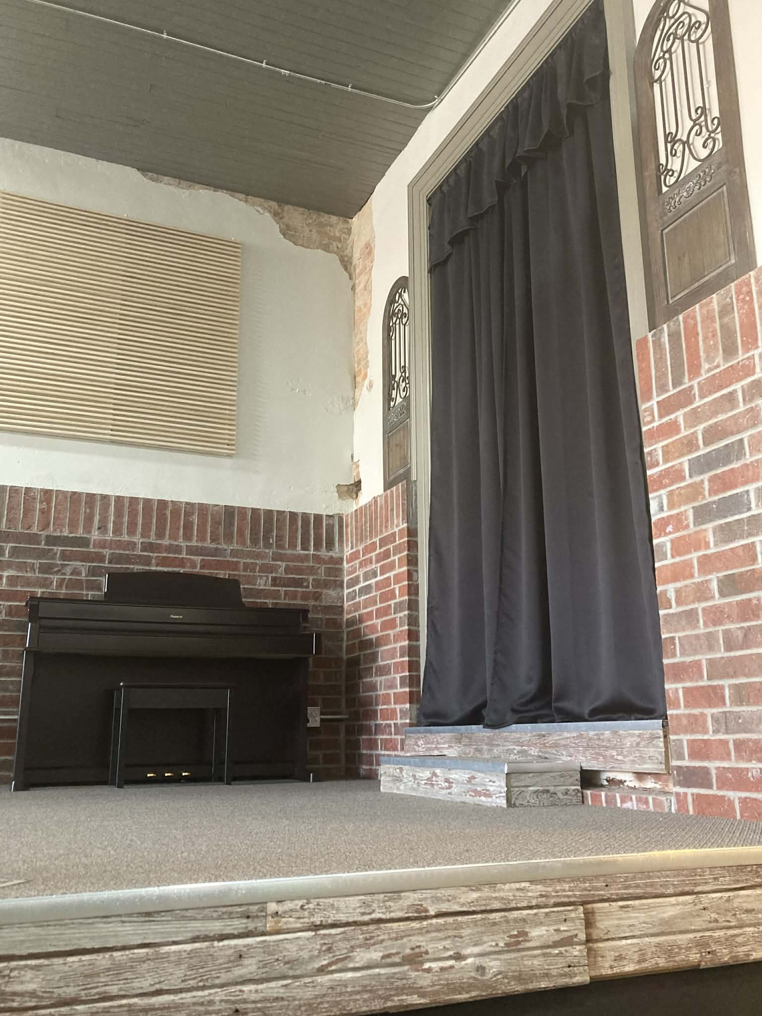 A stage with a black piano against a brick wall and a black curtain on the right. The wall has a fragment of plaster missing near the ceiling, revealing brick behind it, and a window with beige blinds is visible on the left. The stage is made of weathered wood and carpet.
