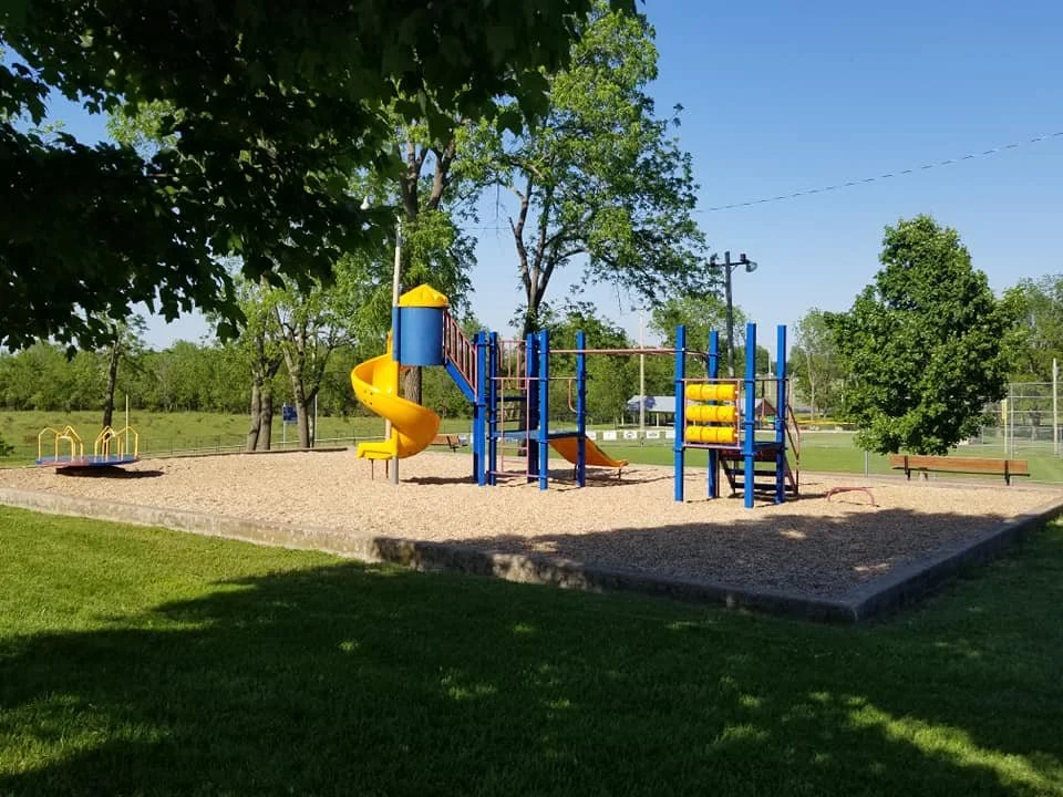 Children's playground with a yellow spiral slide, climbing structures, and swings, surrounded by trees and grass on a sunny day.