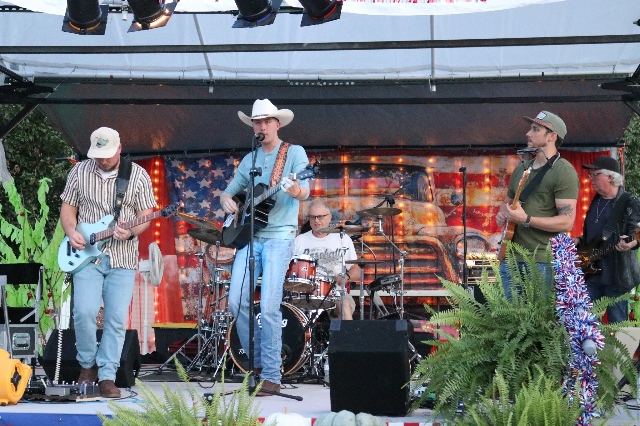 A five-member band performing on an outdoor stage with a patriotic American flag backdrop. The band includes guitarists, a drummer, and a bassist, with the lead singer wearing a cowboy hat.