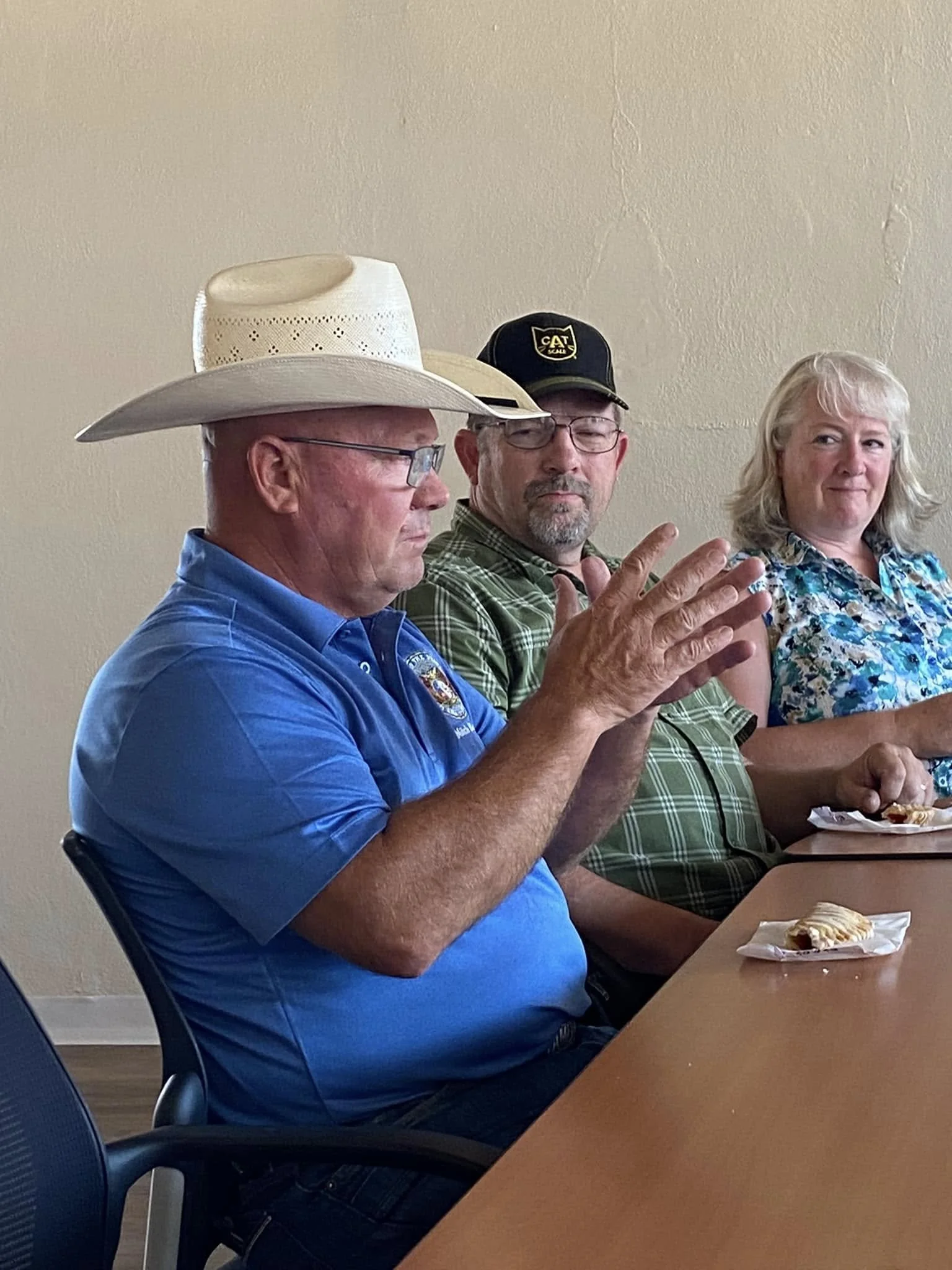 Three people are sitting at a table, one man wearing a cowboy hat and glasses clapping, another man with a black 'CAT' cap and glasses, and a woman with curly blonde hair in a floral shirt. There are pastries on the table.