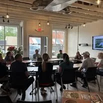 Group of people seated around tables in a modern, well-lit room with large windows, listening to a speaker at the front.
