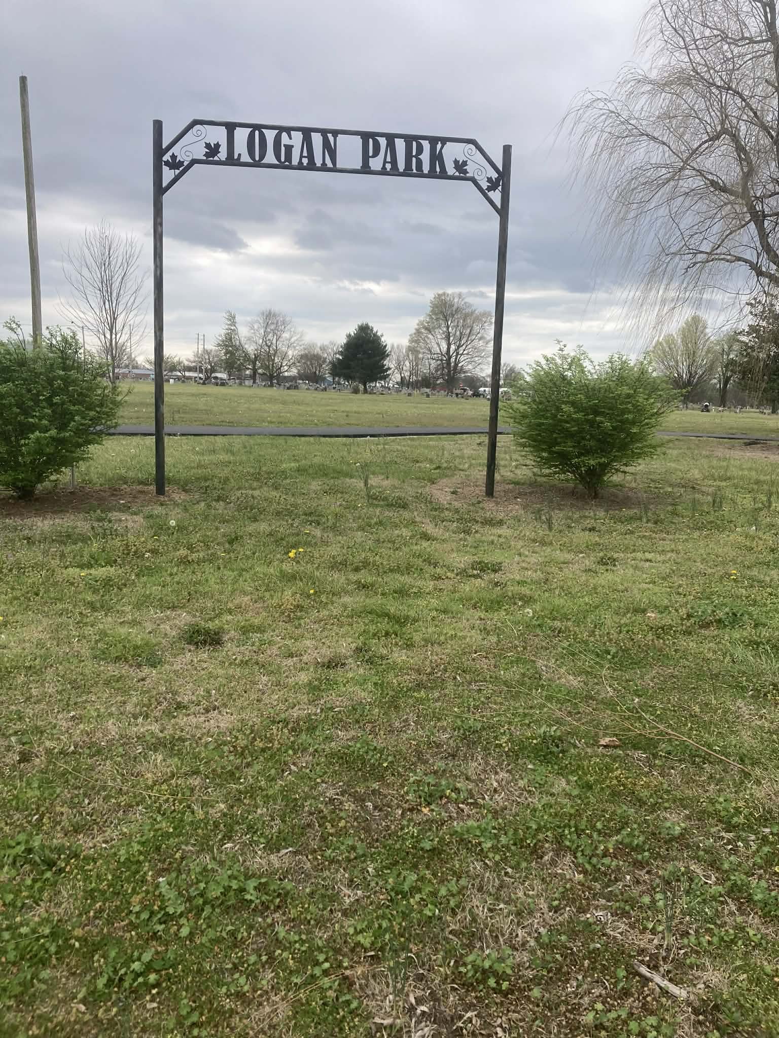 Entrance sign for Logan Park with grass and bushes in the foreground, cloudy sky, and trees in the background.