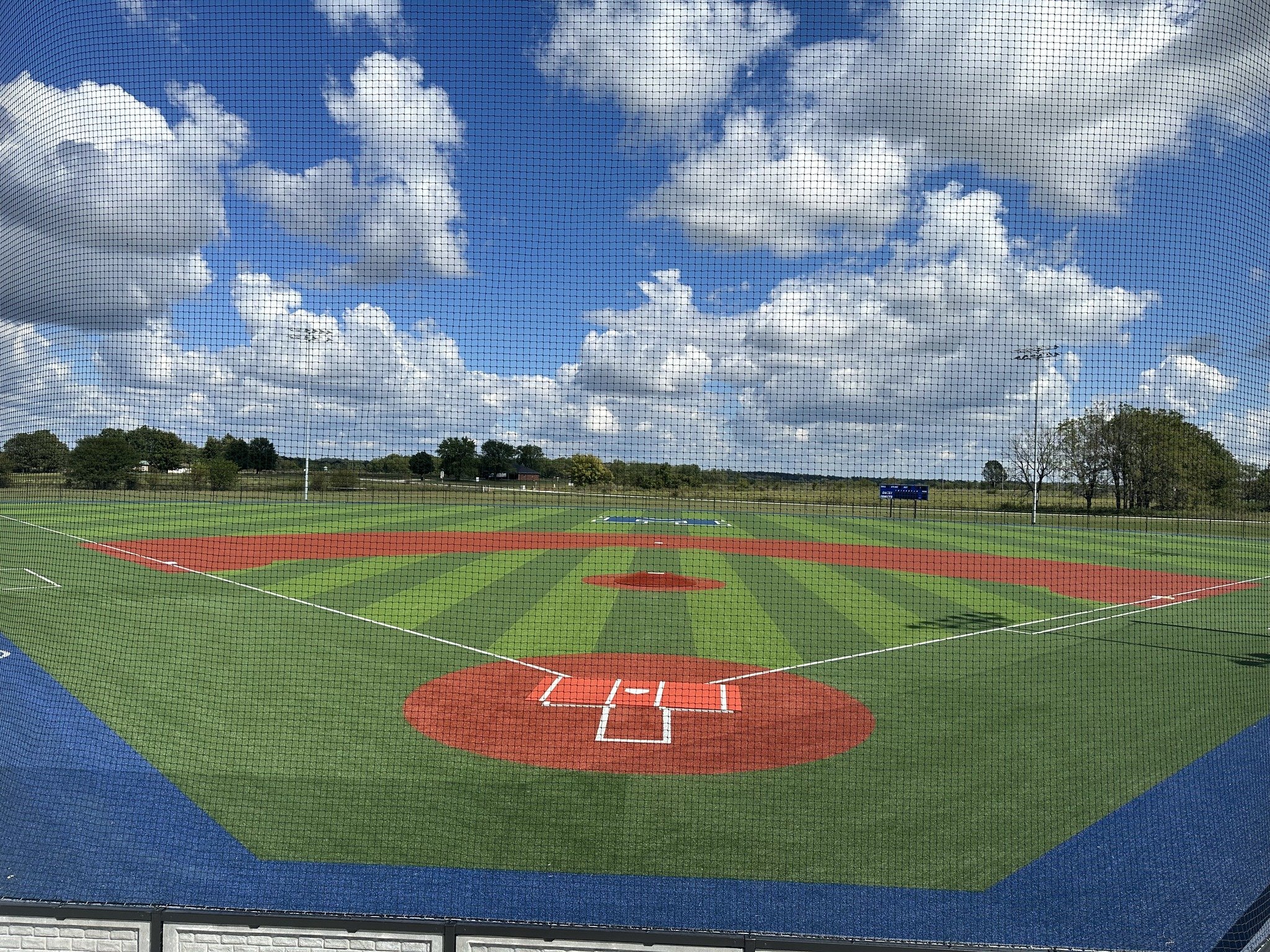 View of a baseball field seen through a chain-link fence, with a partly cloudy sky and green trees in the background.