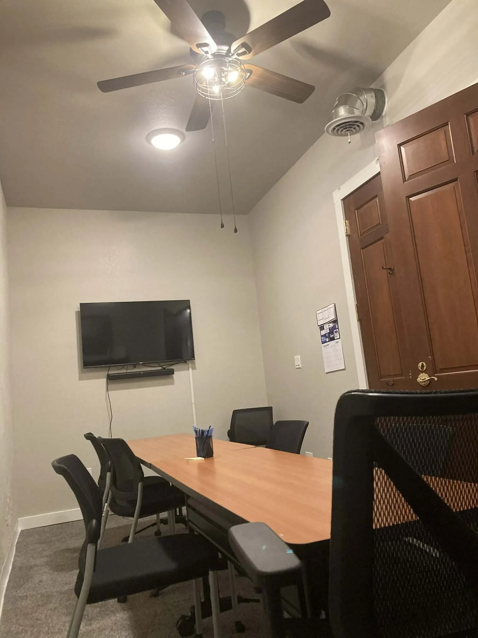 Conference room with a wooden table, black chairs, wall-mounted TV, ceiling fan, ceiling light, clipboard on the wall, and a wooden door.