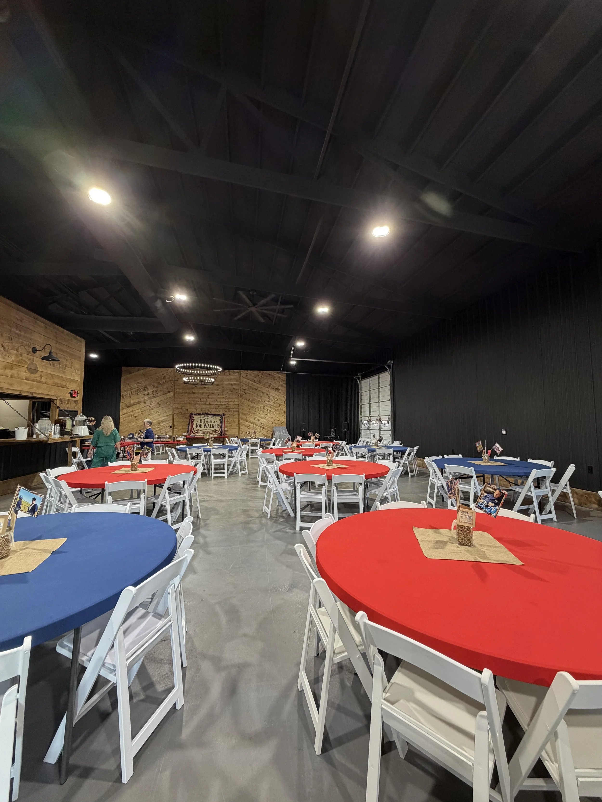 Interior of a restaurant with round tables covered in red, blue, and black tablecloths, white chairs, and a rustic wood wall at the back. There are people, a service counter, and decorative lighting fixtures.