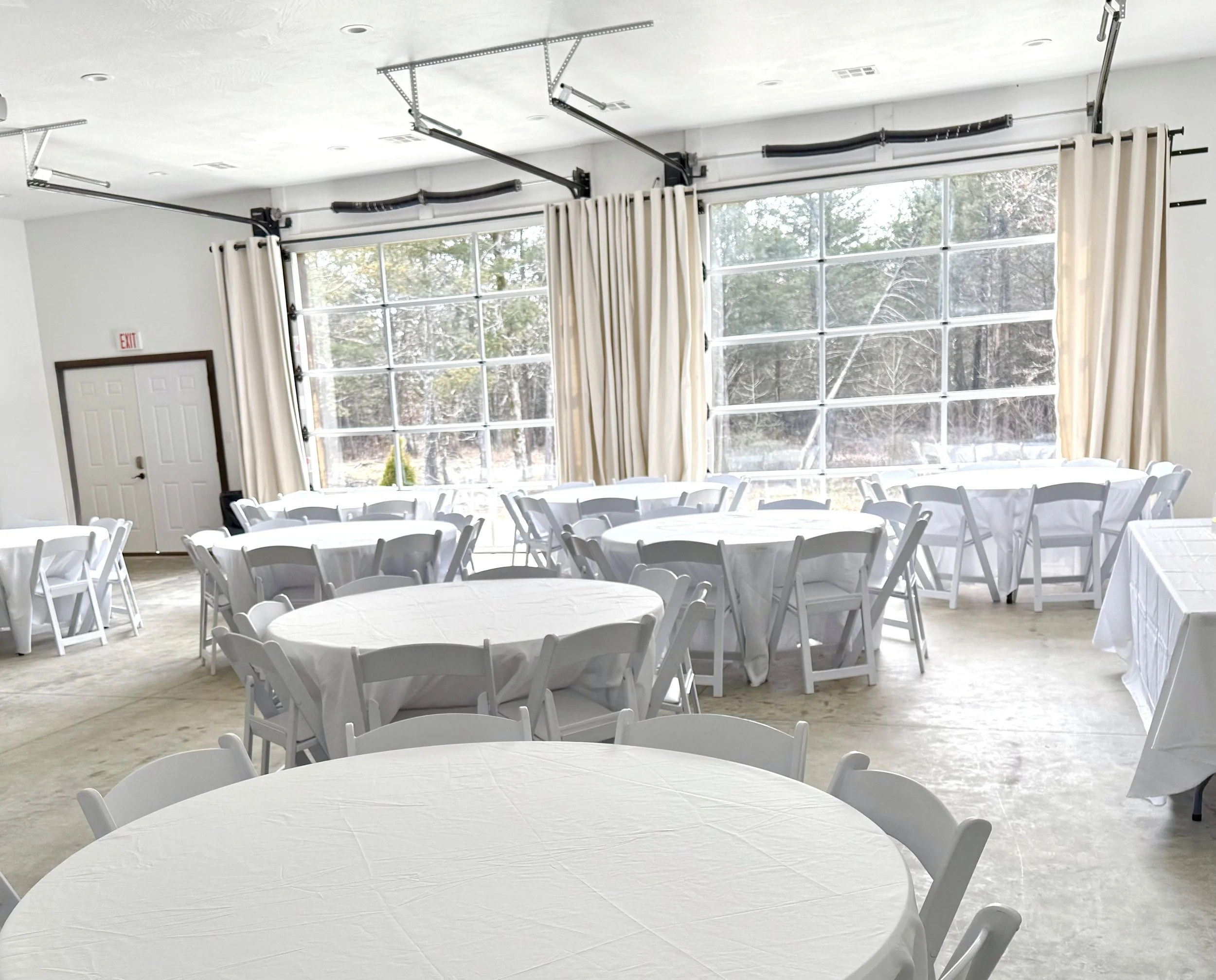 A banquet hall with round tables covered in white tablecloths and white chairs. Large windows with cream curtains allow natural light to fill the room, overlooking a snowy outdoor scene.