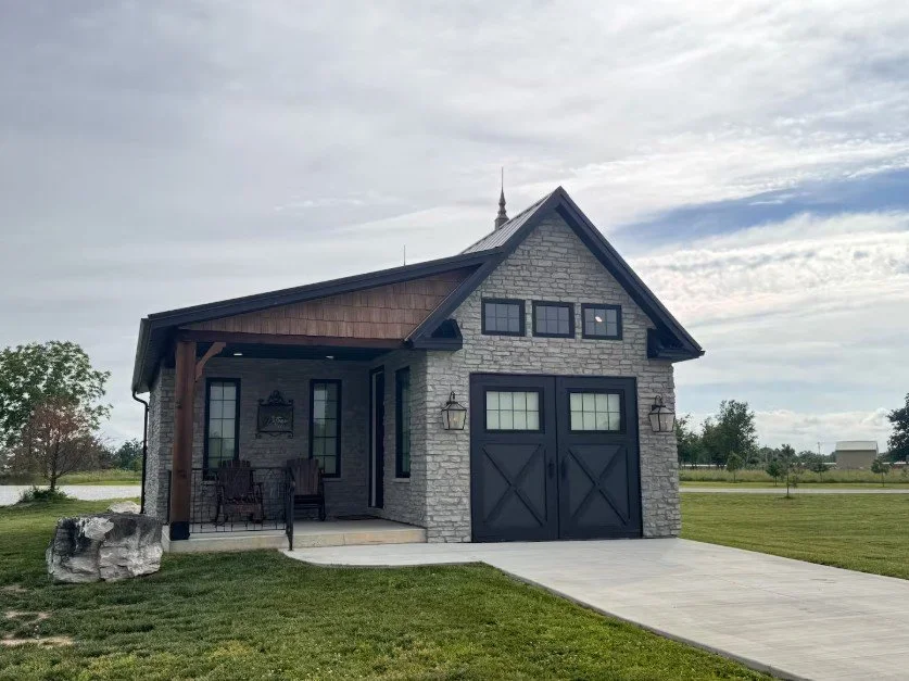 A modern detached garage with a stone facade, dark barn-style doors, and small window above the doors, situated in a grassy area with a driveway and a small porch with outdoor seating.