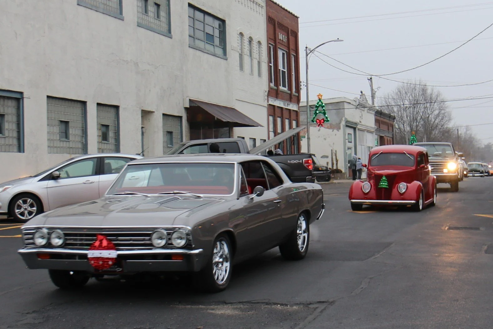 Street scene with decorated vintage cars, including a gray car and a red car with Christmas tree ornaments, and a line of vehicles in the background on a gray winter day.