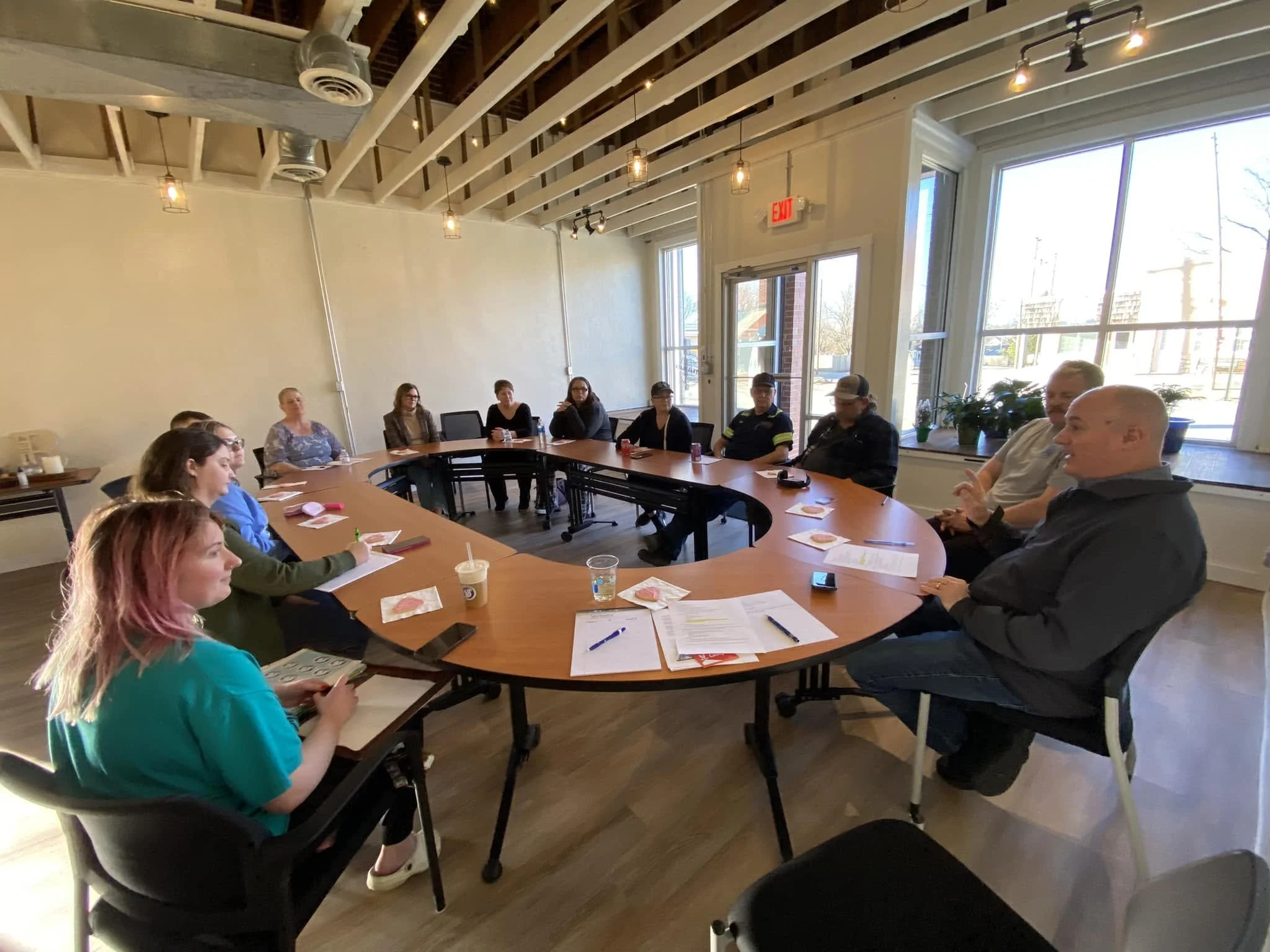 People seated around a large oval conference table during a meeting inside a room with large windows and exposed ceiling beams.