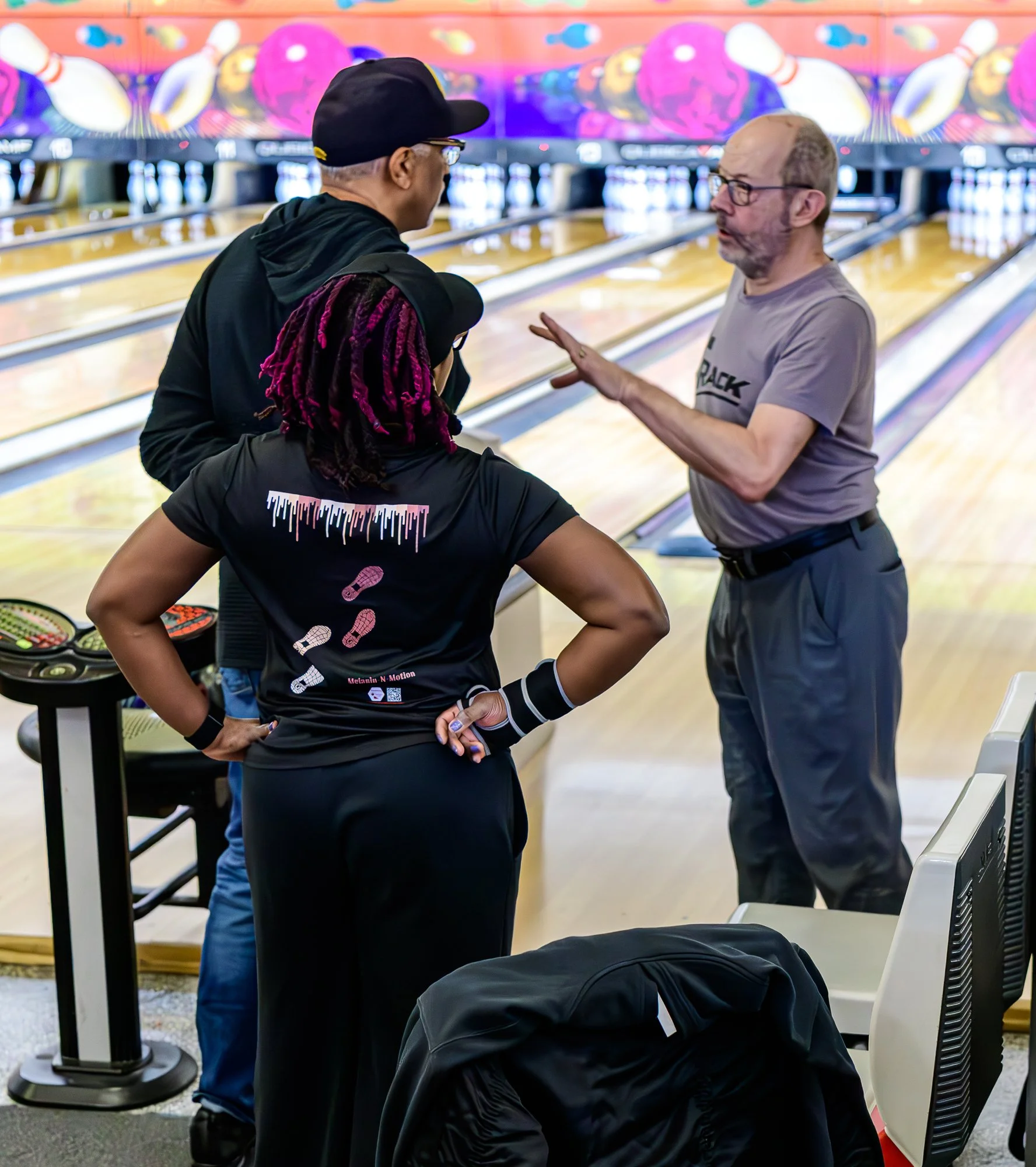 A group of people at a bowling alley, including a woman with colorful dreadlocks, a man in a gray t-shirt, and another man in a black jacket and cap, engaged in a conversation.