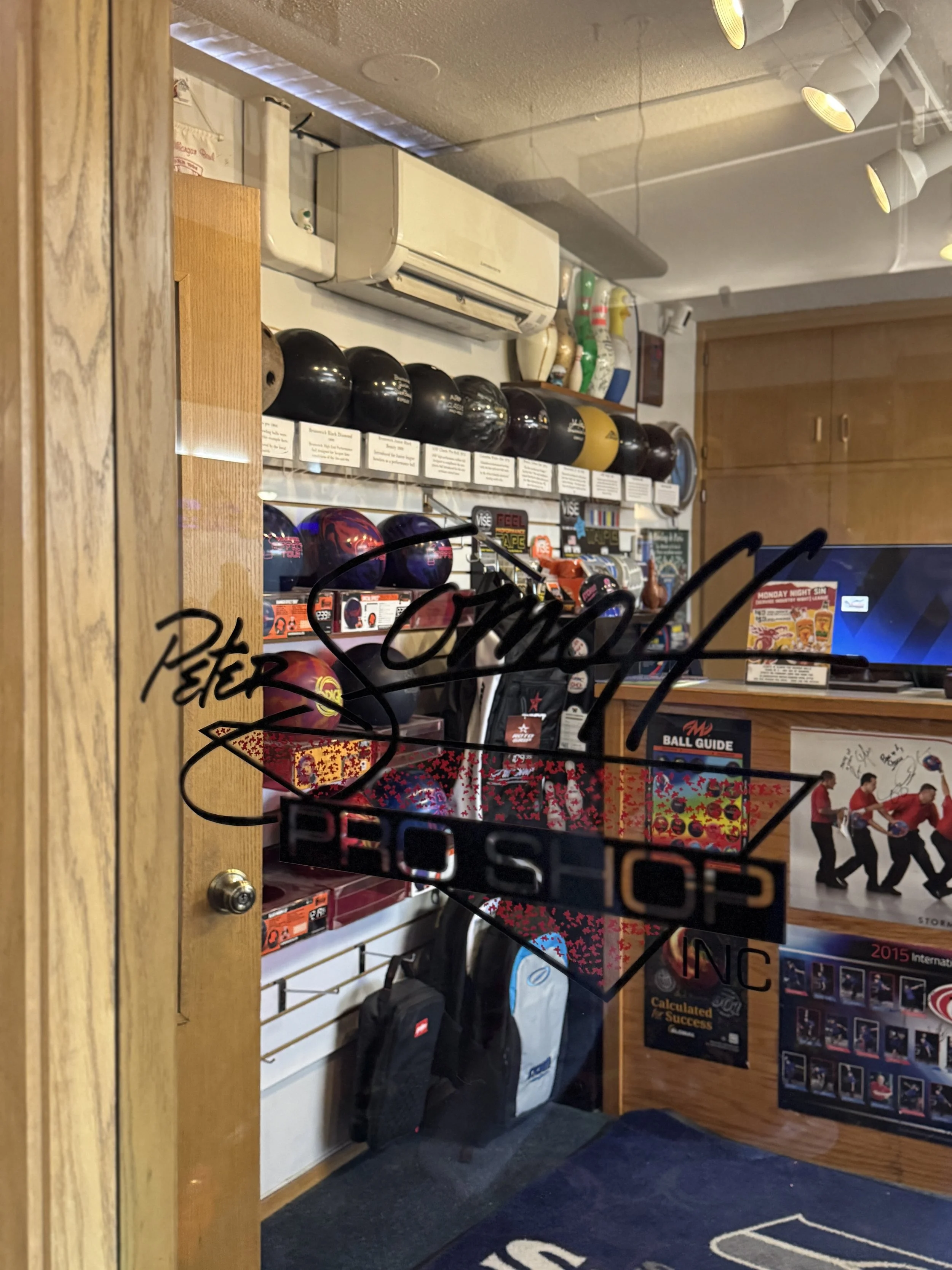 View through a glass door showing bowling equipment, including several bowling balls on shelves, bowling bags, and posters in a bowling alley or pro shop.