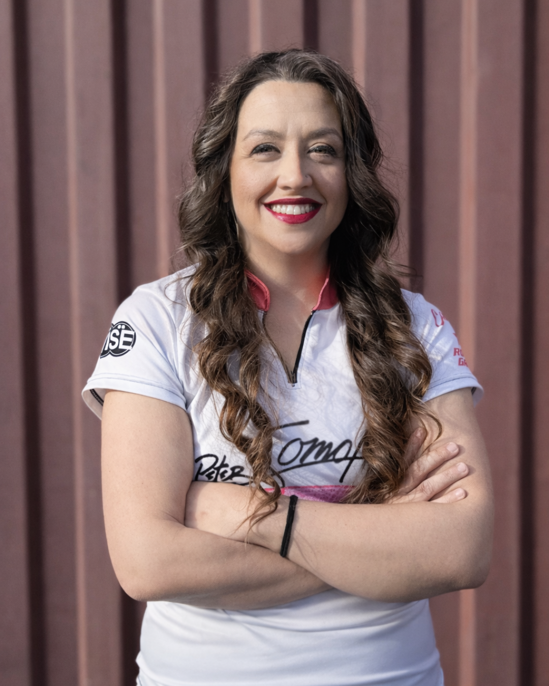 A woman with long curly brown hair smiling and crossing her arms, wearing a white sports shirt with various logos and signatures, standing in front of a red-brown vertical panel background.