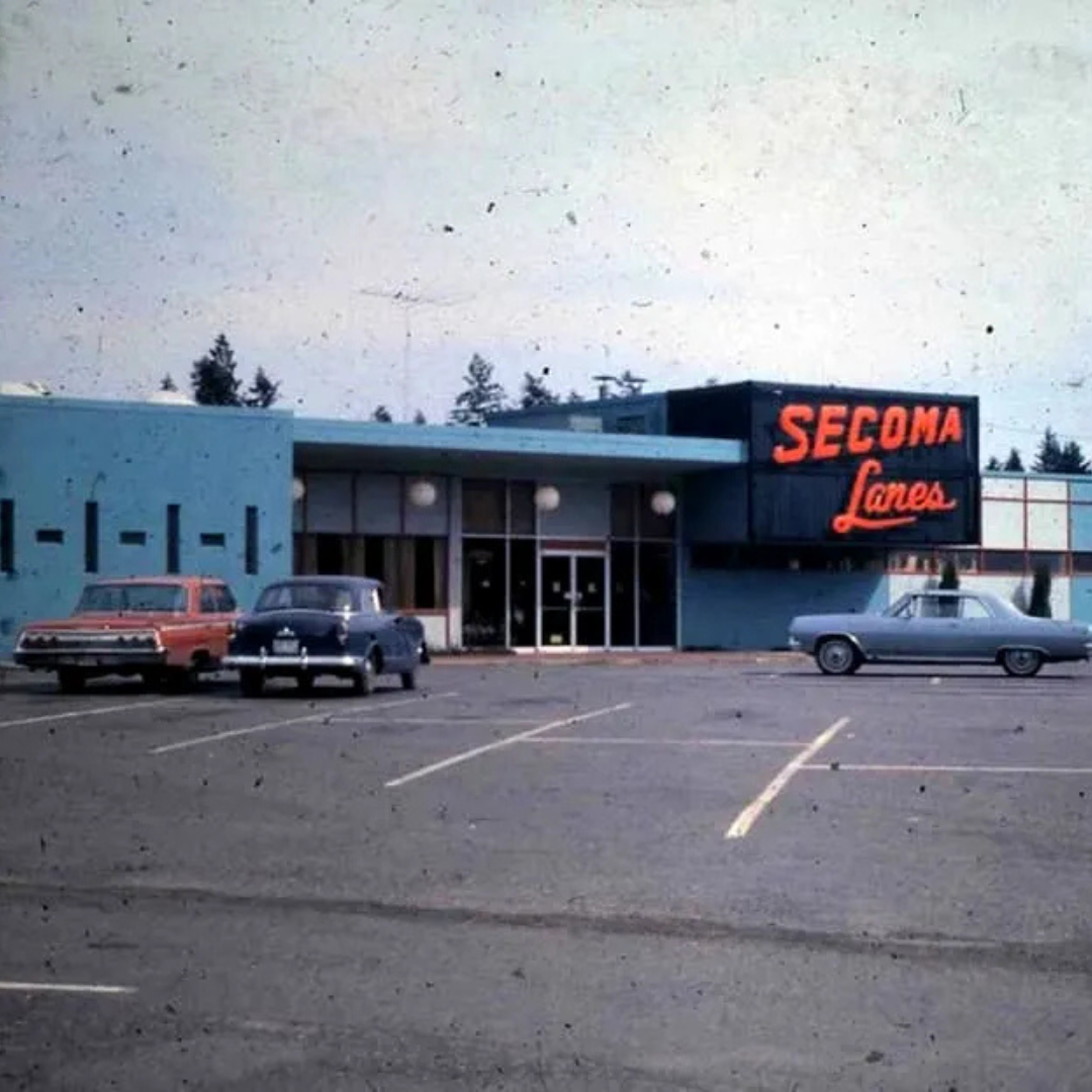 Old parking lot with three vintage cars parked in front of a retro-style building, which has a sign reading 'Secoma Lanes' in bright red neon letters.