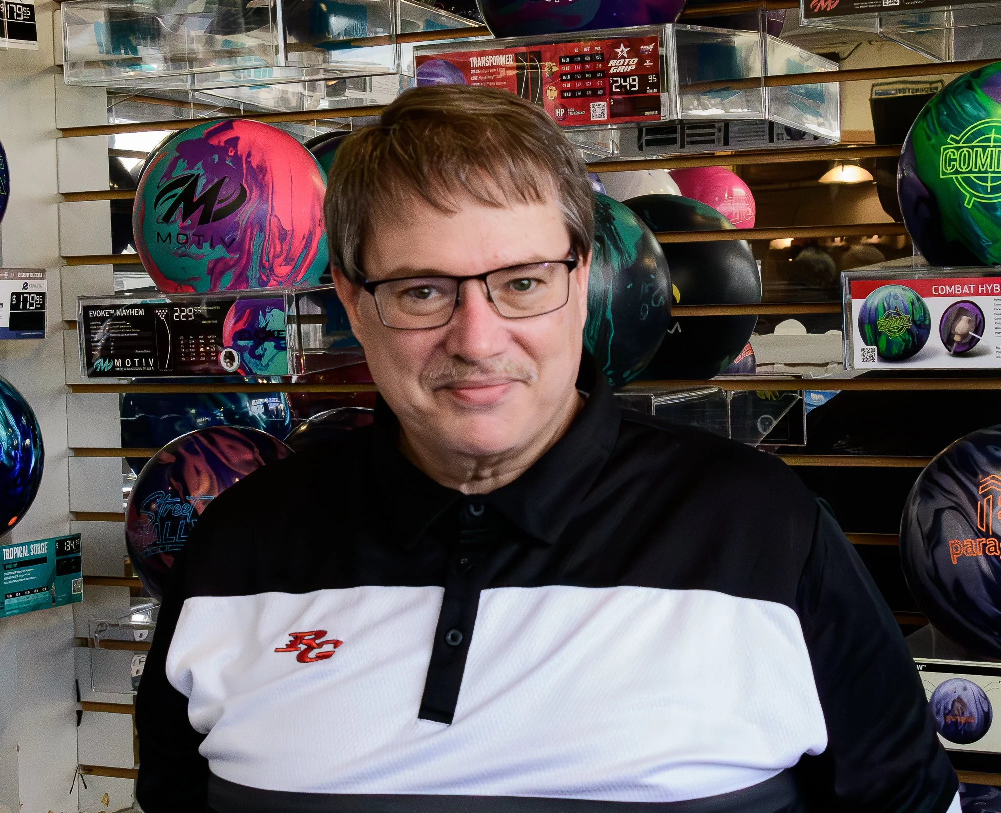 A man with glasses and short brown hair, wearing a black and white polo shirt, standing in front of a display of colorful bowling balls in a store.