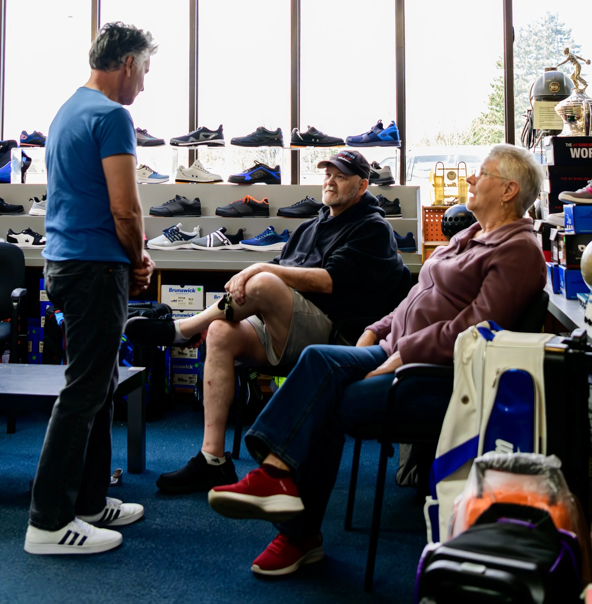 A man and woman sitting in a shoe store talking to a man standing in front of them. The seated man has a bandaged knee with a brace and is wearing shorts and a hoodie. The seated woman is dressed in a hoodie and jeans. The background shows shelves of